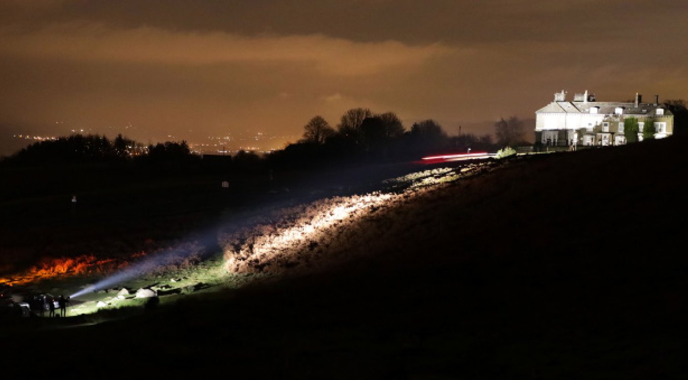 Nighttime scene of a sloped hillside with a white building at the top right, illuminated rocks and dirt on the hill, and light trails from a passing vehicle on the right side; a small group of people with flashlights near the bottom left.