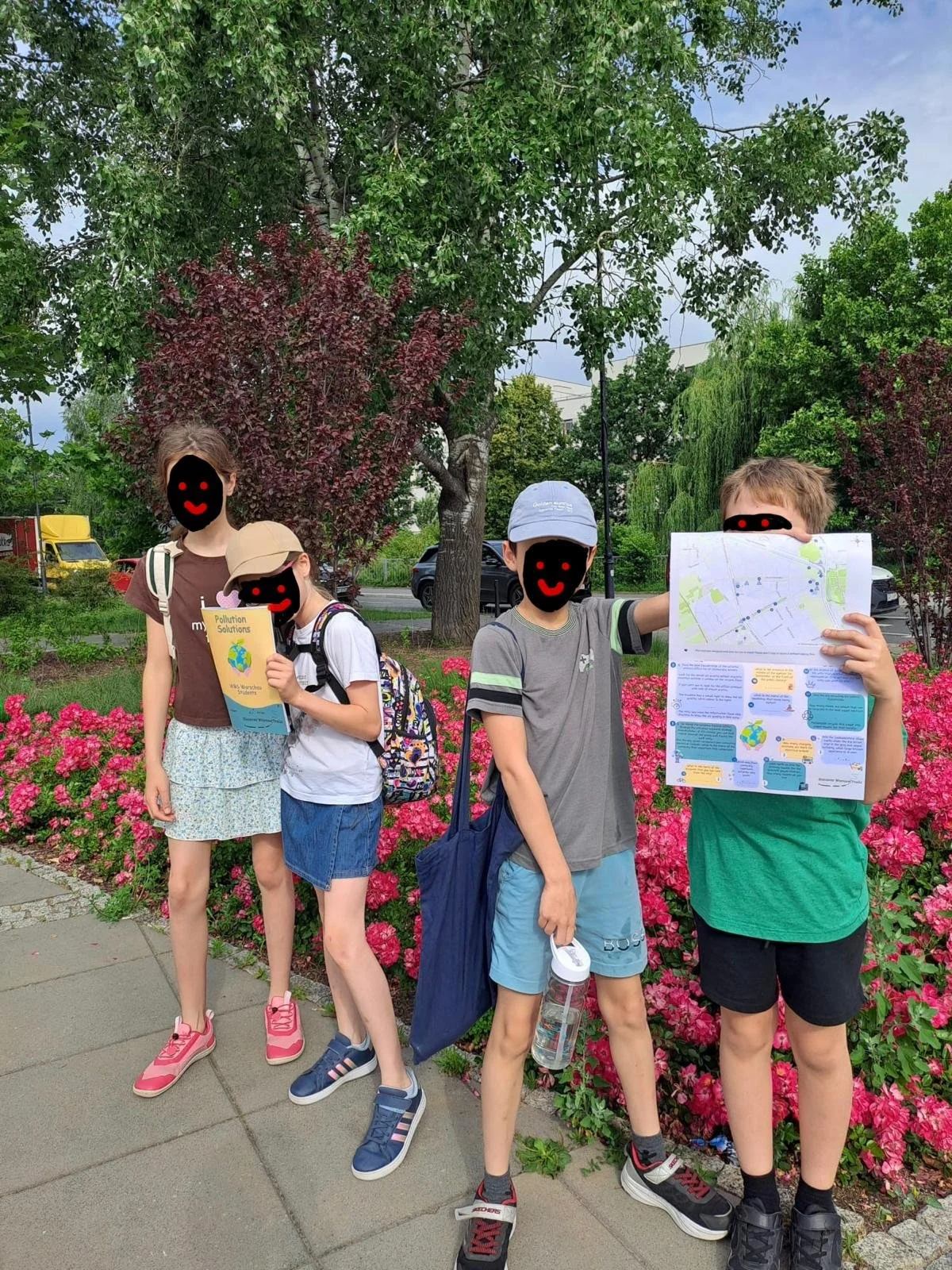 Four children standing outdoors on a sidewalk in front of a flower bed with pink flowers, trees, and cars in the background. One girl is holding a piece of paper with a map, and the other girl and two boys are holding papers or books related to pollution solutions, wearing backpacks and hats.