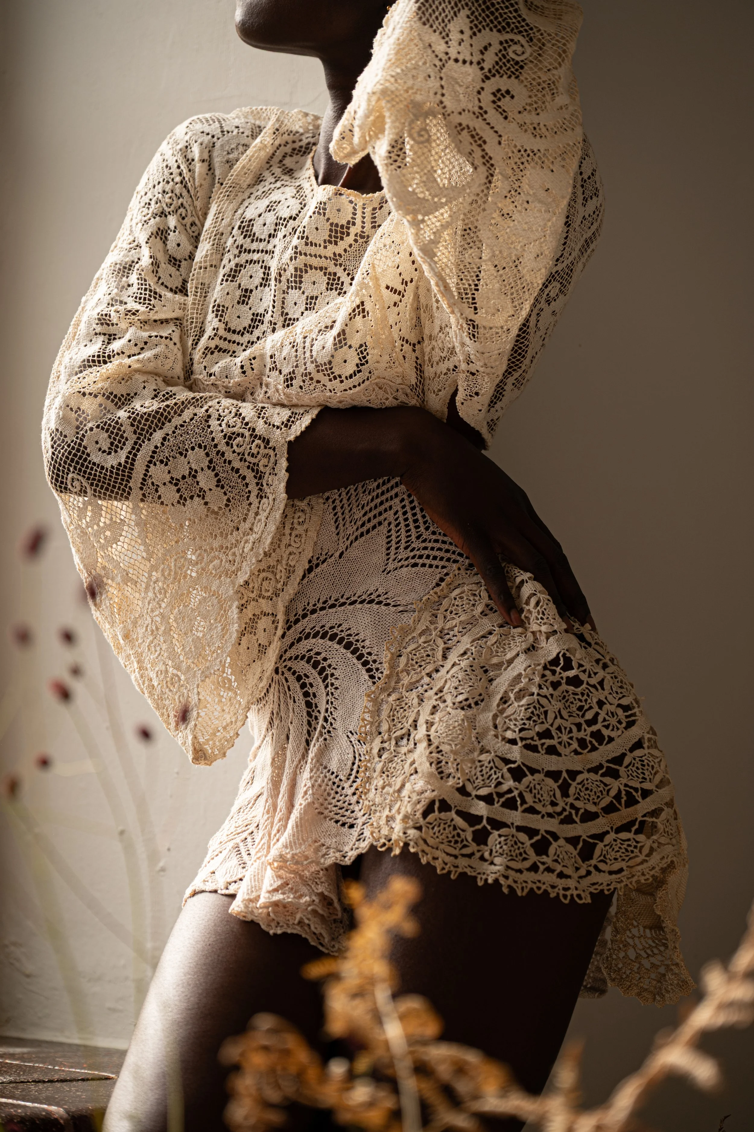 Close-up of a woman wearing vintage cream-colored lace dress with intricate patterns, posing with one arm crossed and her hand resting on her thigh, against a plain wall with floral elements in the foreground.