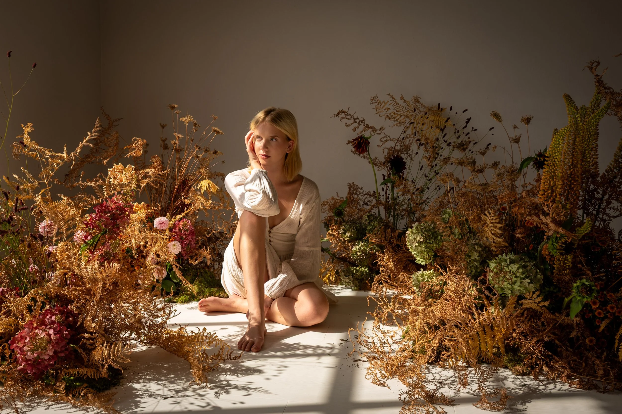 A woman sitting on the floor amid dried brown and green plants and flowers, wearing a light-colored dress, with one hand on her face, looking to the side, against a plain wall background.
