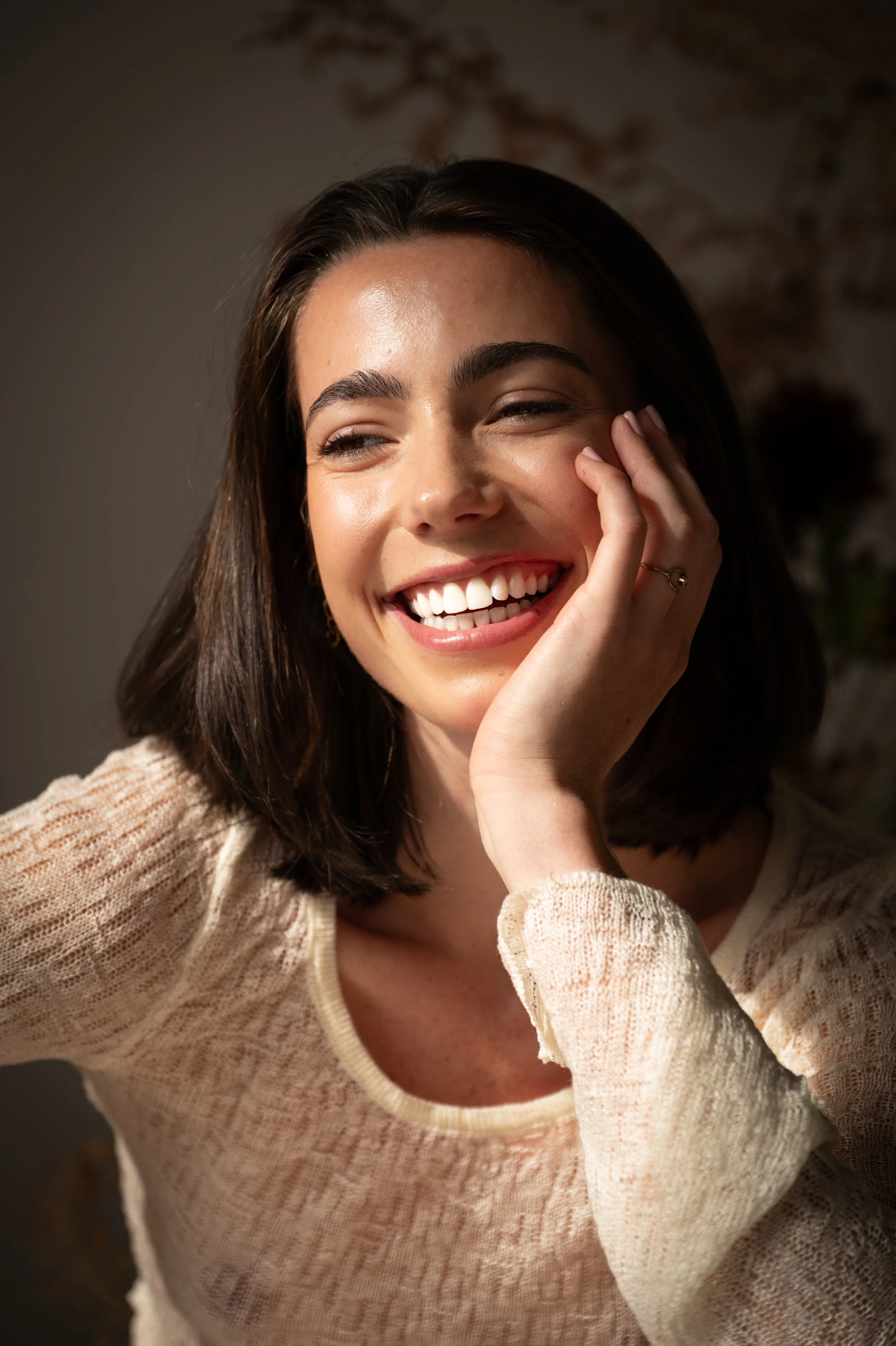 A young woman with dark brown hair and fair skin smiling and touching her face with her left hand, wearing a cream-colored sweater, in a warm, softly lit setting.