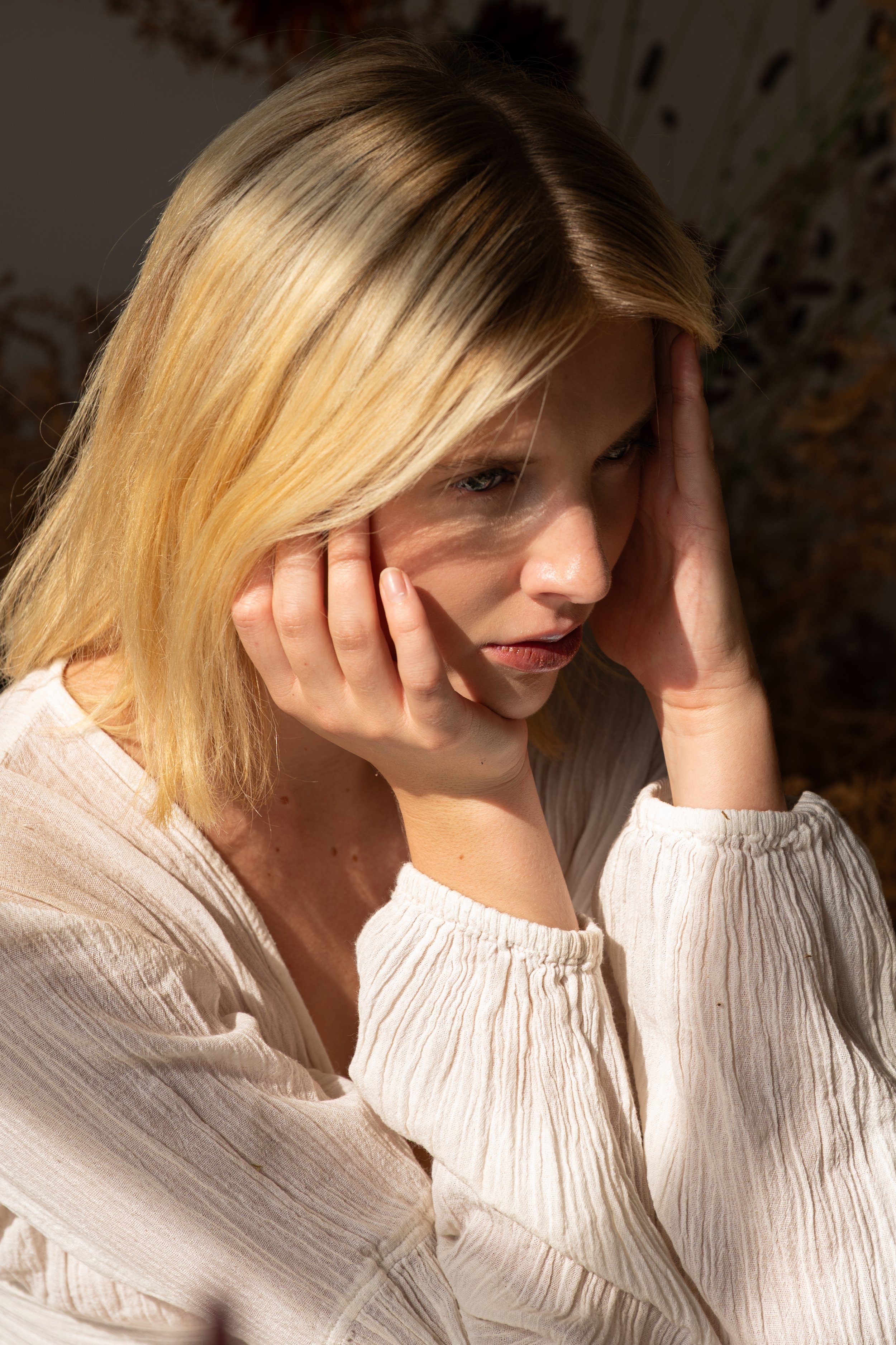 Young woman with blonde hair, holding her head with both hands, appearing distressed or deep in thought, wearing a light-colored, textured long sleeve top.