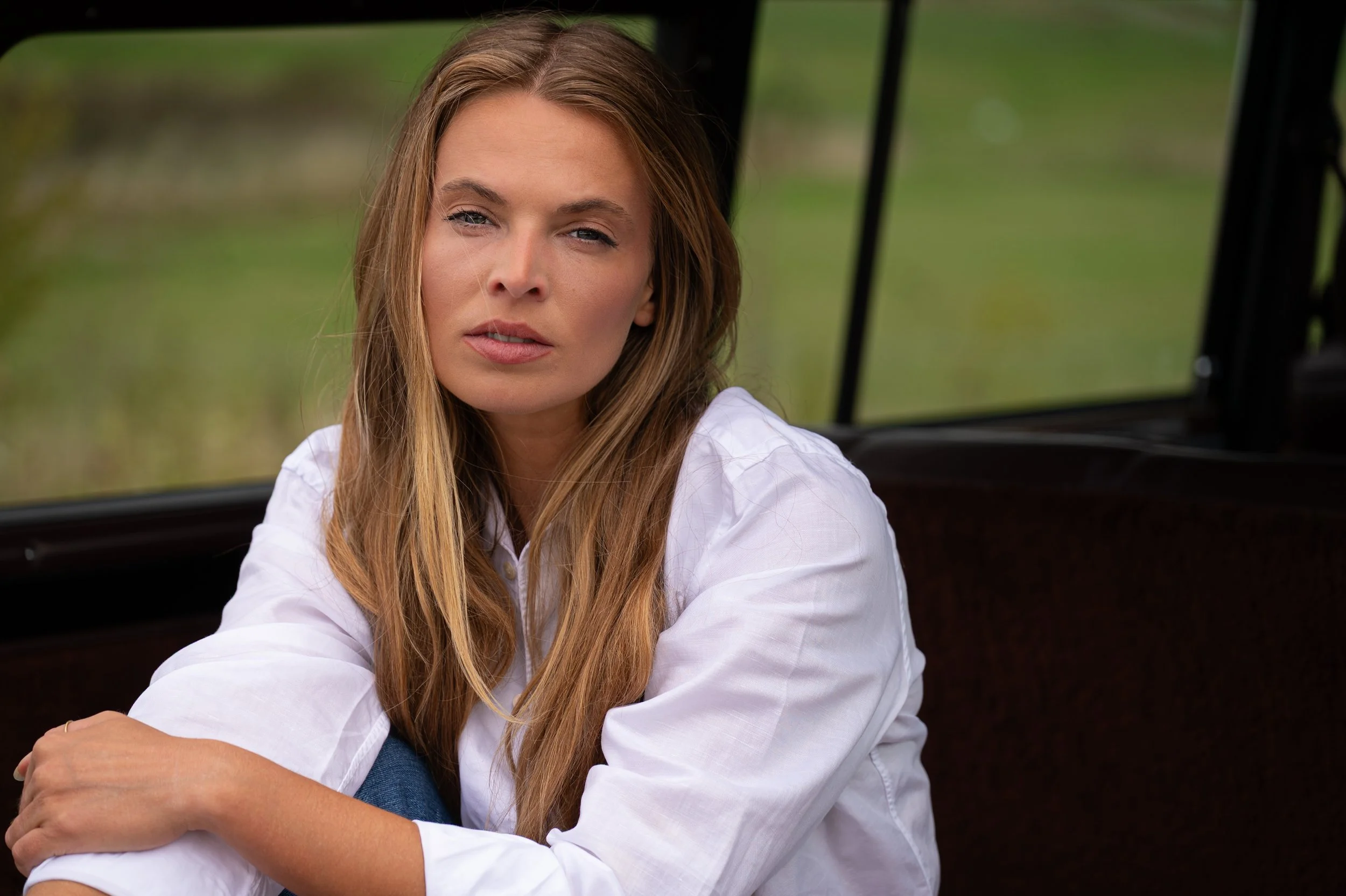 A woman with long, light brown hair and a white shirt is sitting inside a vehicle, looking directly at the camera with a serious expression. The background shows greenery outside the vehicle.