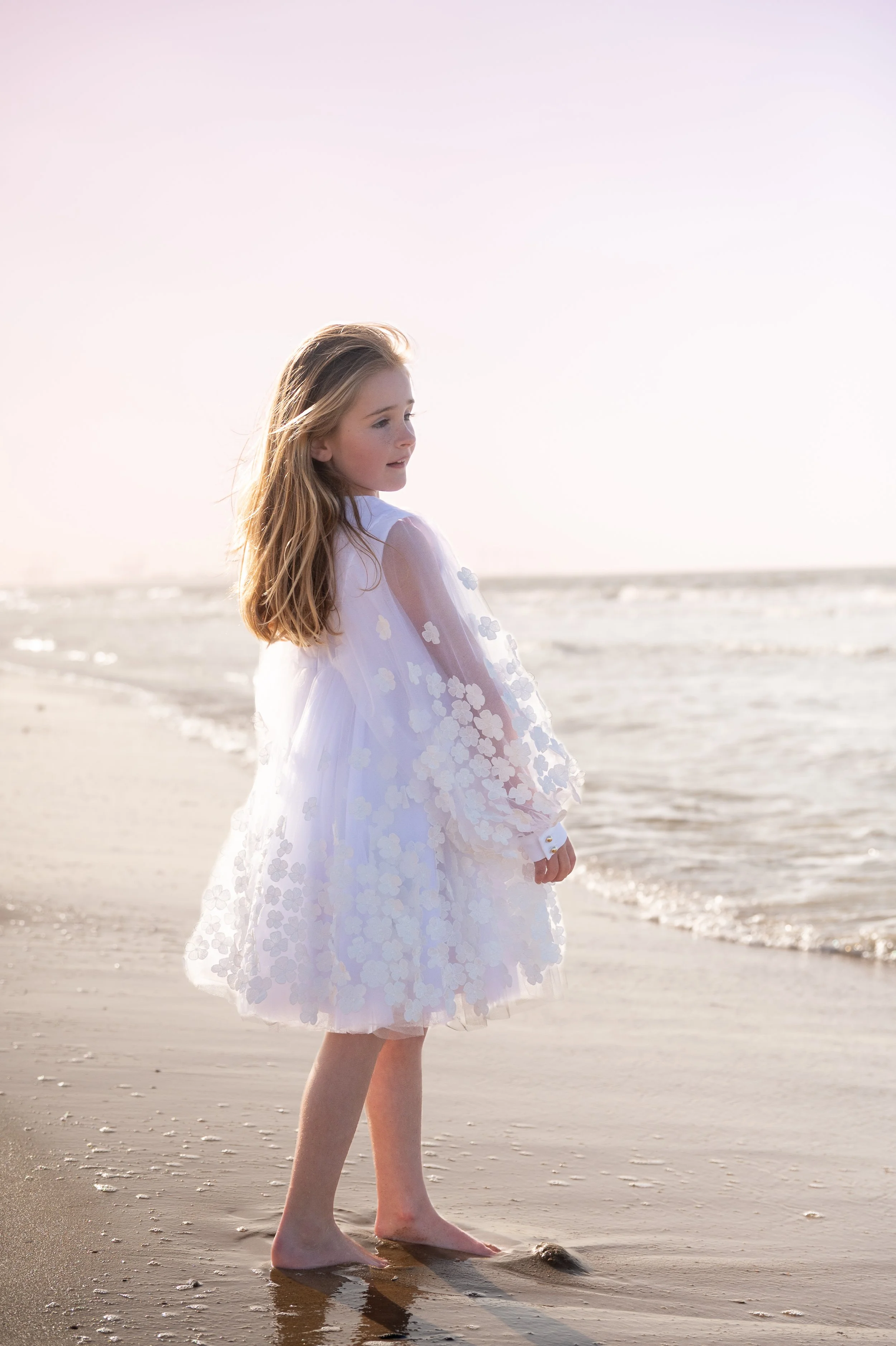 A young girl in a white dress with floral details standing barefoot on the beach near the water at sunset or sunrise.