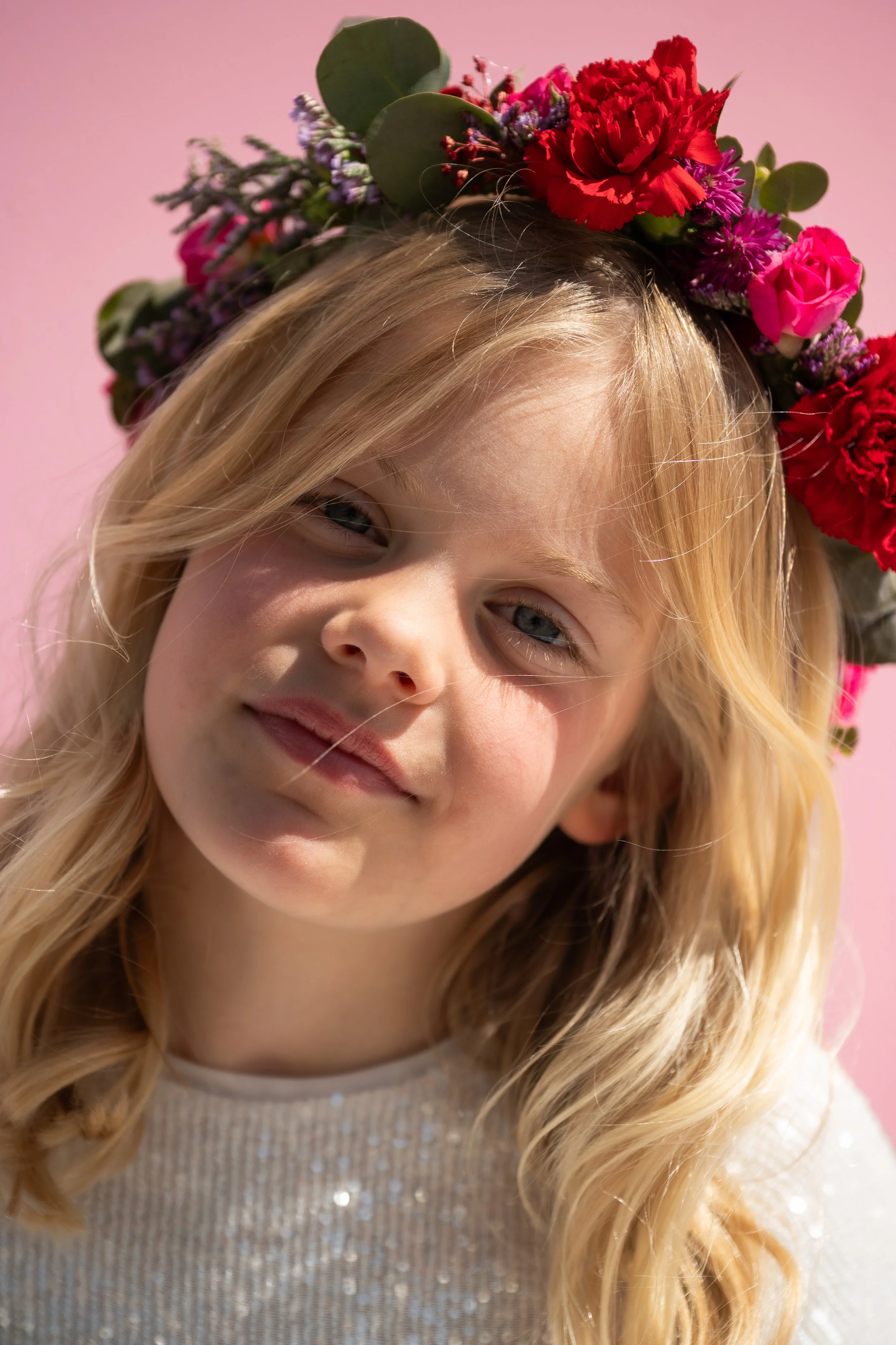 A young girl with blonde hair and blue eyes wearing a flower crown with red and purple flowers against a pink background.