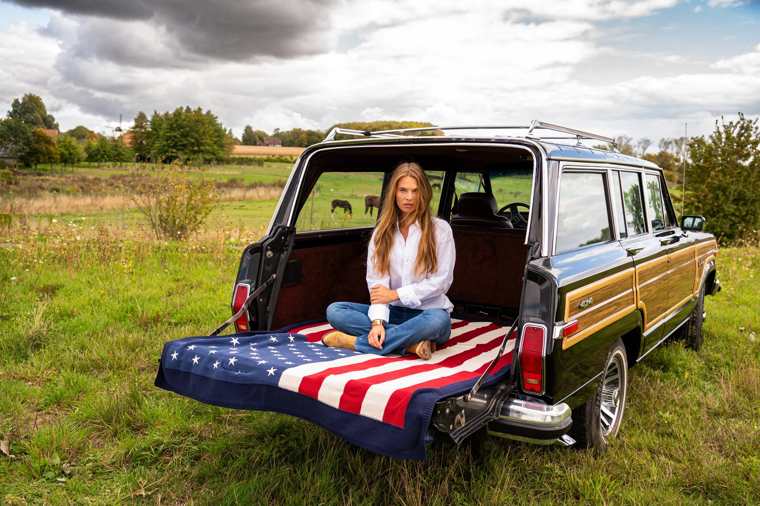 A woman sitting cross-legged in the back of a vintage black station wagon with wood paneling, which has an American flag blanket in the trunk, in a grassy field with trees and horses in the background.
