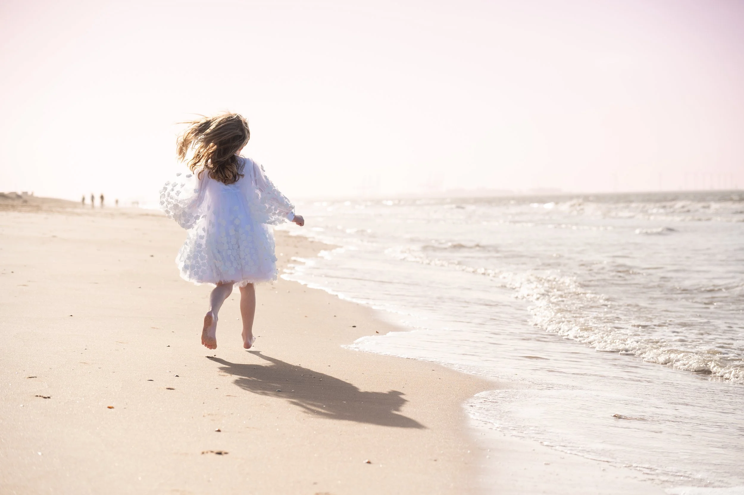 A young girl in a white dress running along the sandy beach near the ocean at sunset or sunrise.