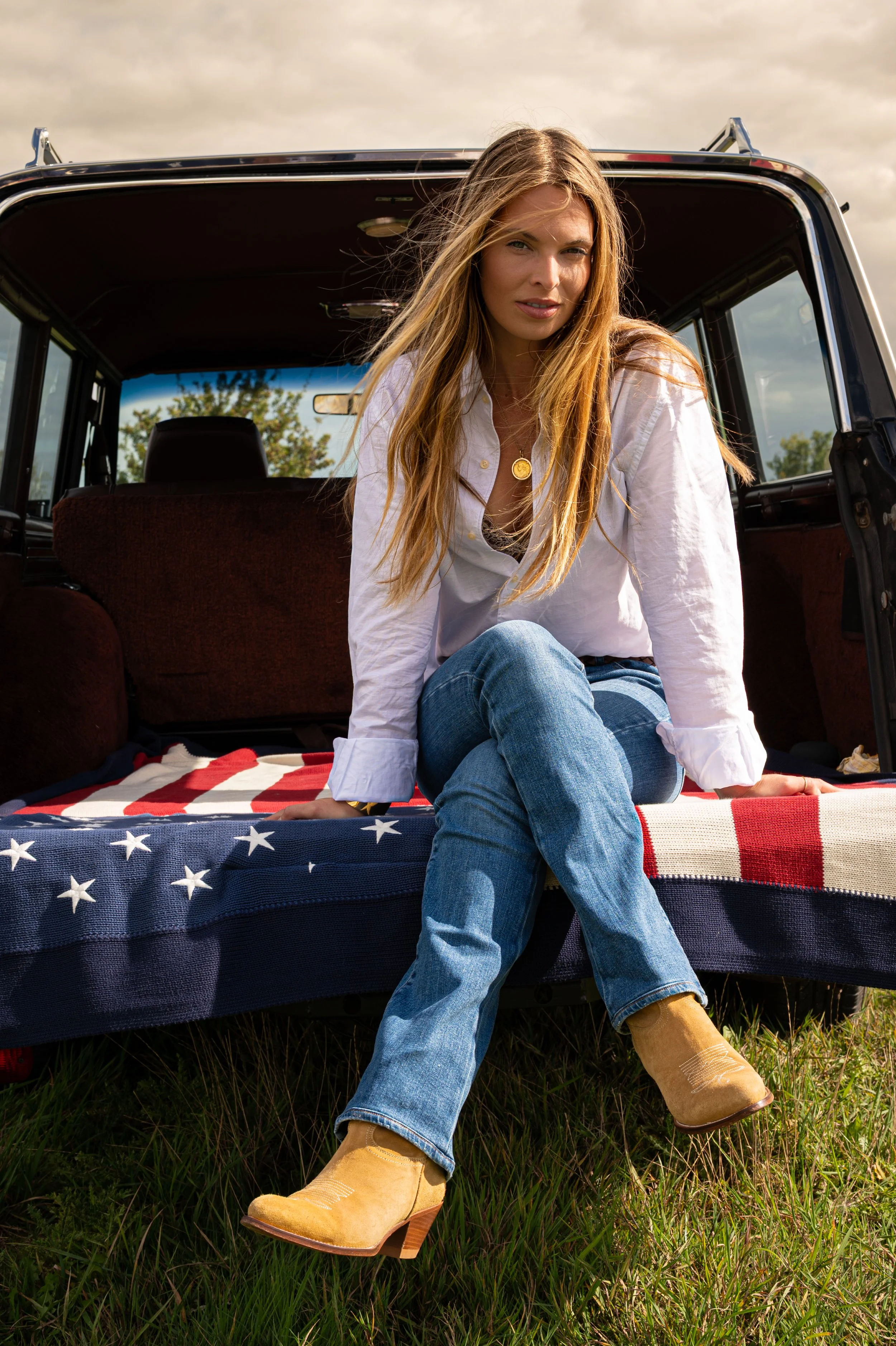 A woman with long blonde hair sitting on the back of a vintage vehicle, wearing a white shirt, jeans, and tan cowboy boots, with a blanket featuring an American flag design underneath her.