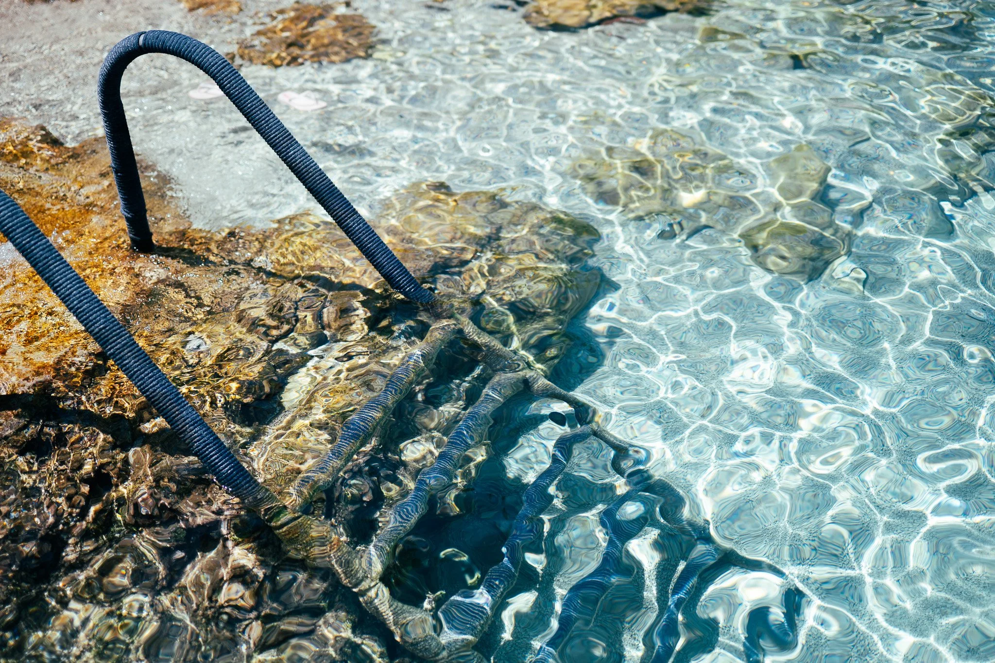 Ocean water with clear ripples, rocky shore with metal pool ladder partially submerged.