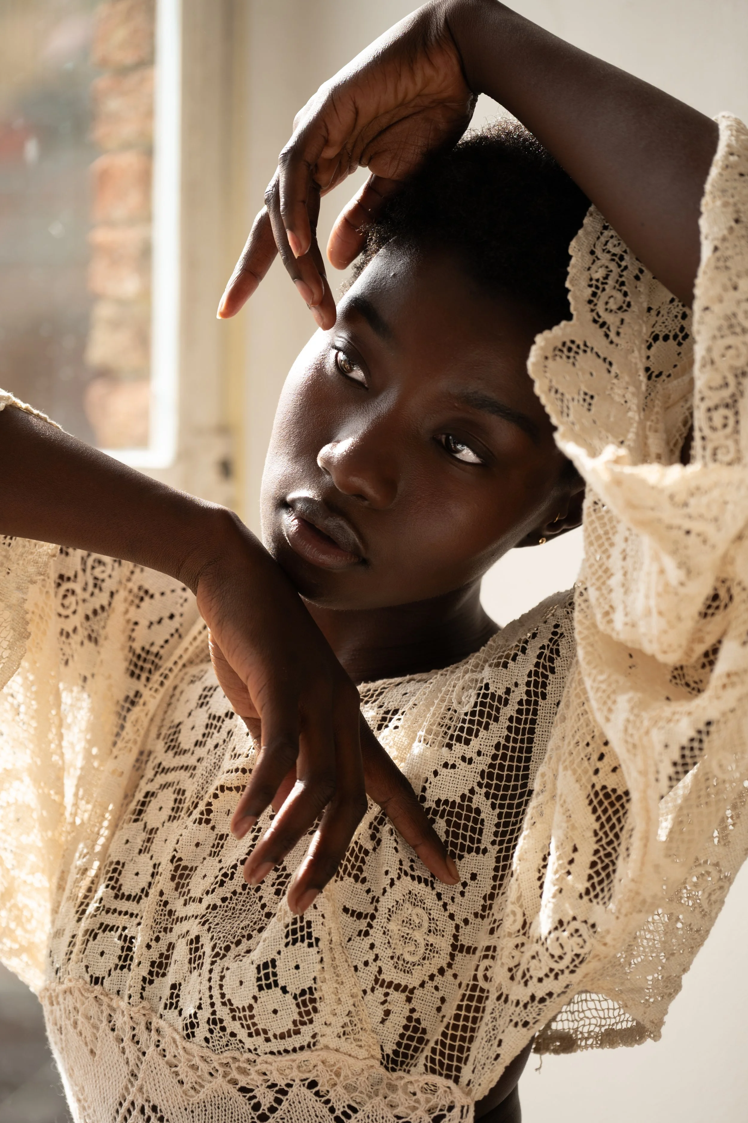 A young woman with short hair and dark skin posed indoors near a window, wearing a detailed cream-colored lace top.