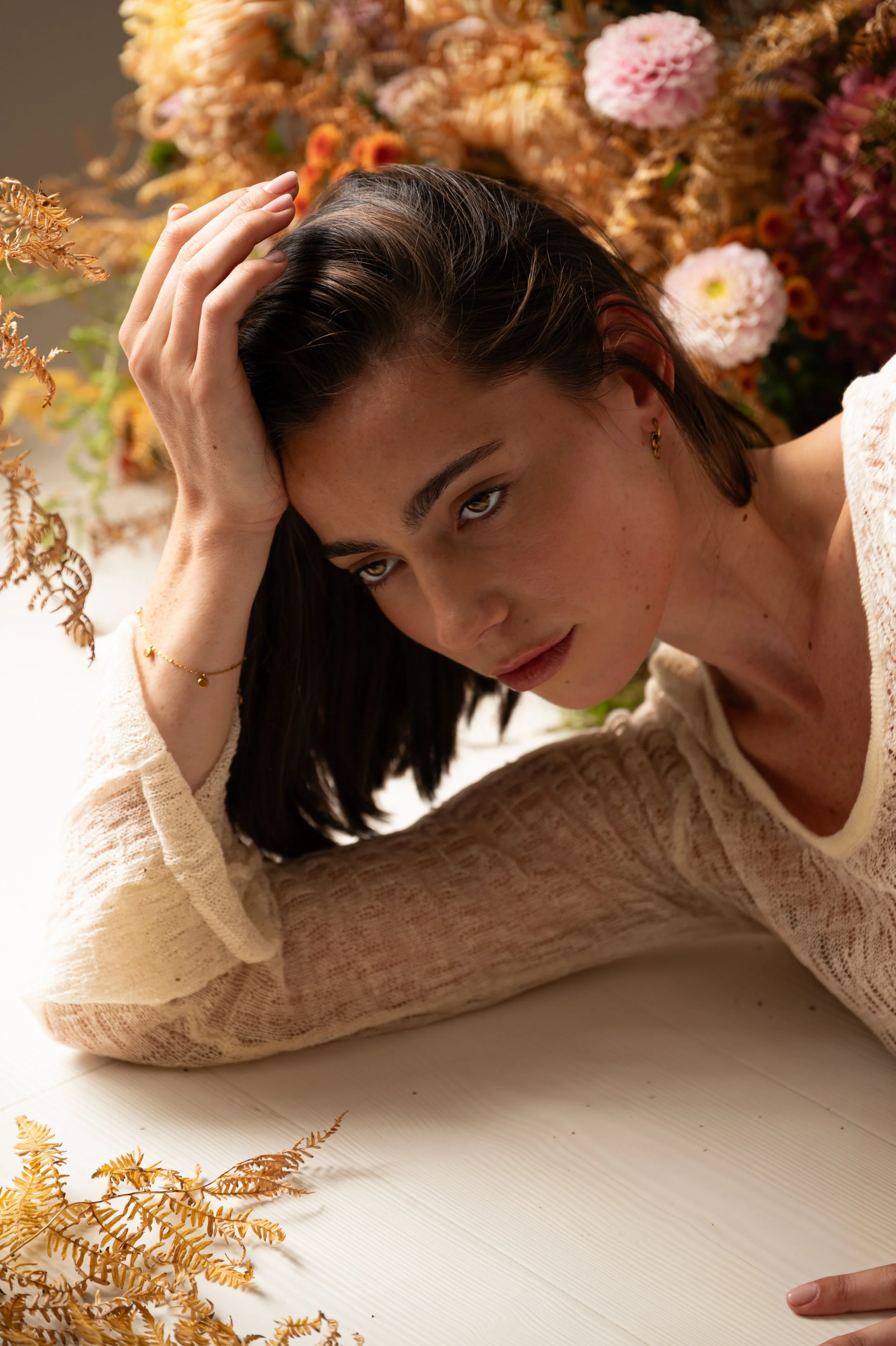 A woman with dark hair and light skin lying on a white wooden floor with her head resting on her left hand. She is wearing a cream-colored, sheer long-sleeve top and gold jewelry. In the background, there are pink and orange flowers and dried foliage.