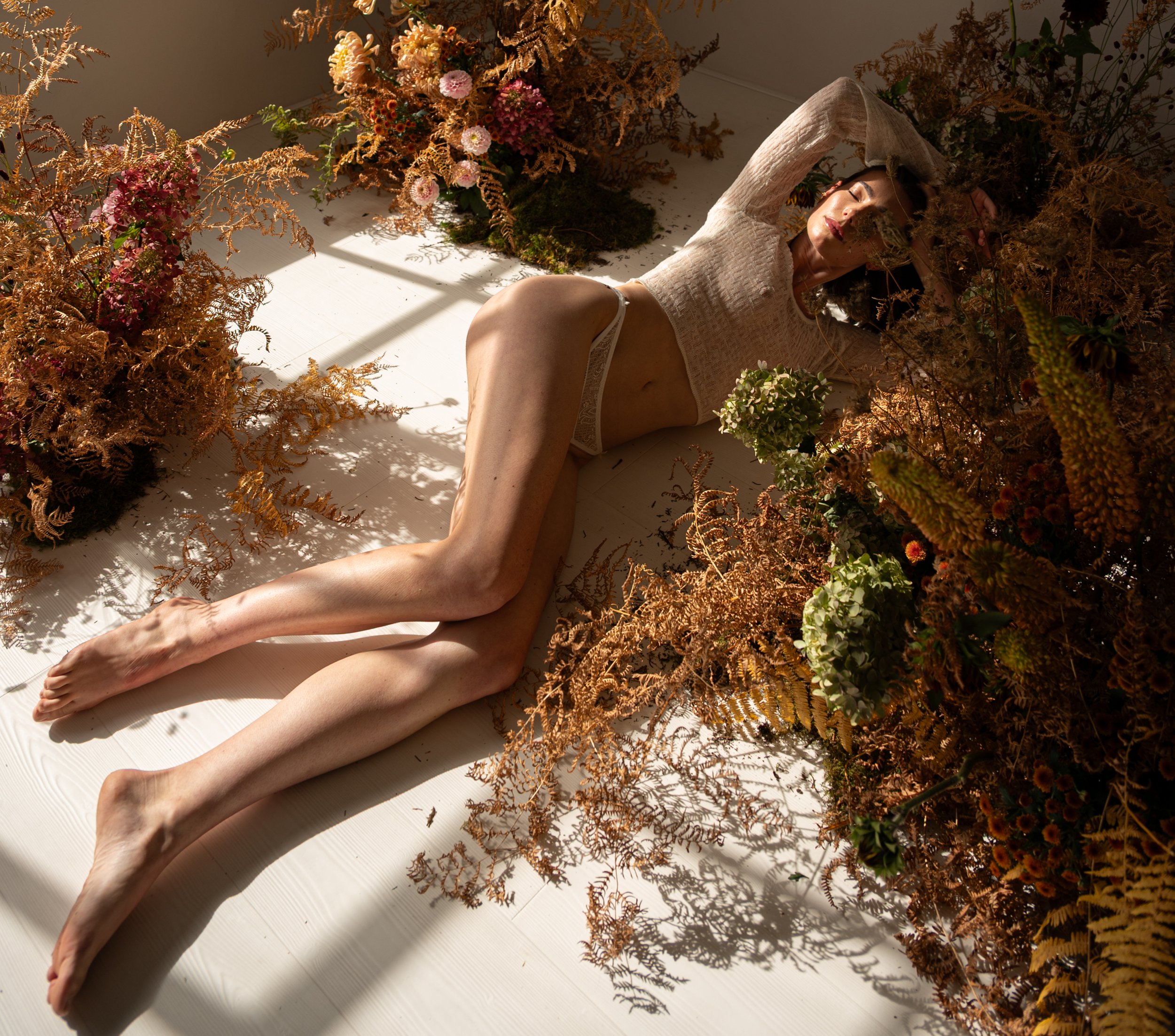 A woman lies on a white floor surrounded by brown and green autumn-themed plants and flowers, with sunlight creating shadows.