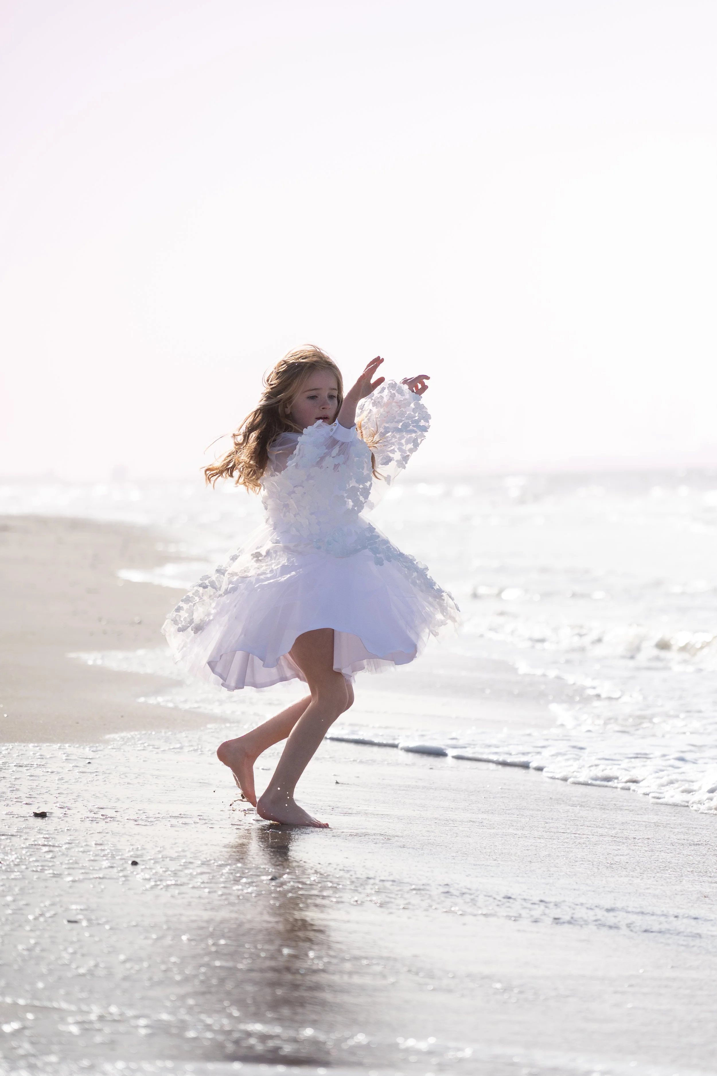 A young girl in a white dress running barefoot on the beach near the shoreline with ocean waves in the background.