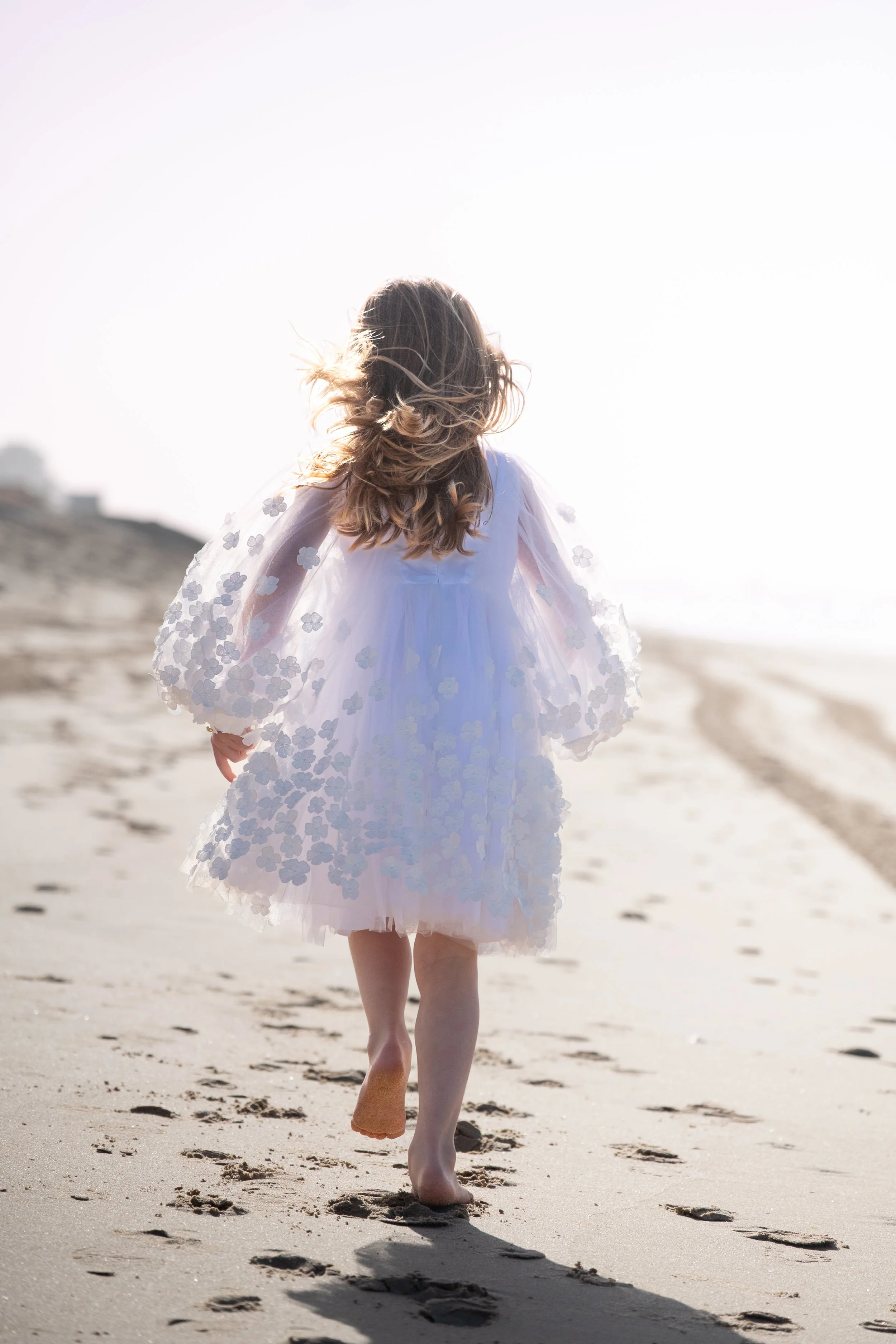 A young girl in a white dress with floral details walking barefoot on a sandy beach at sunset or sunrise.