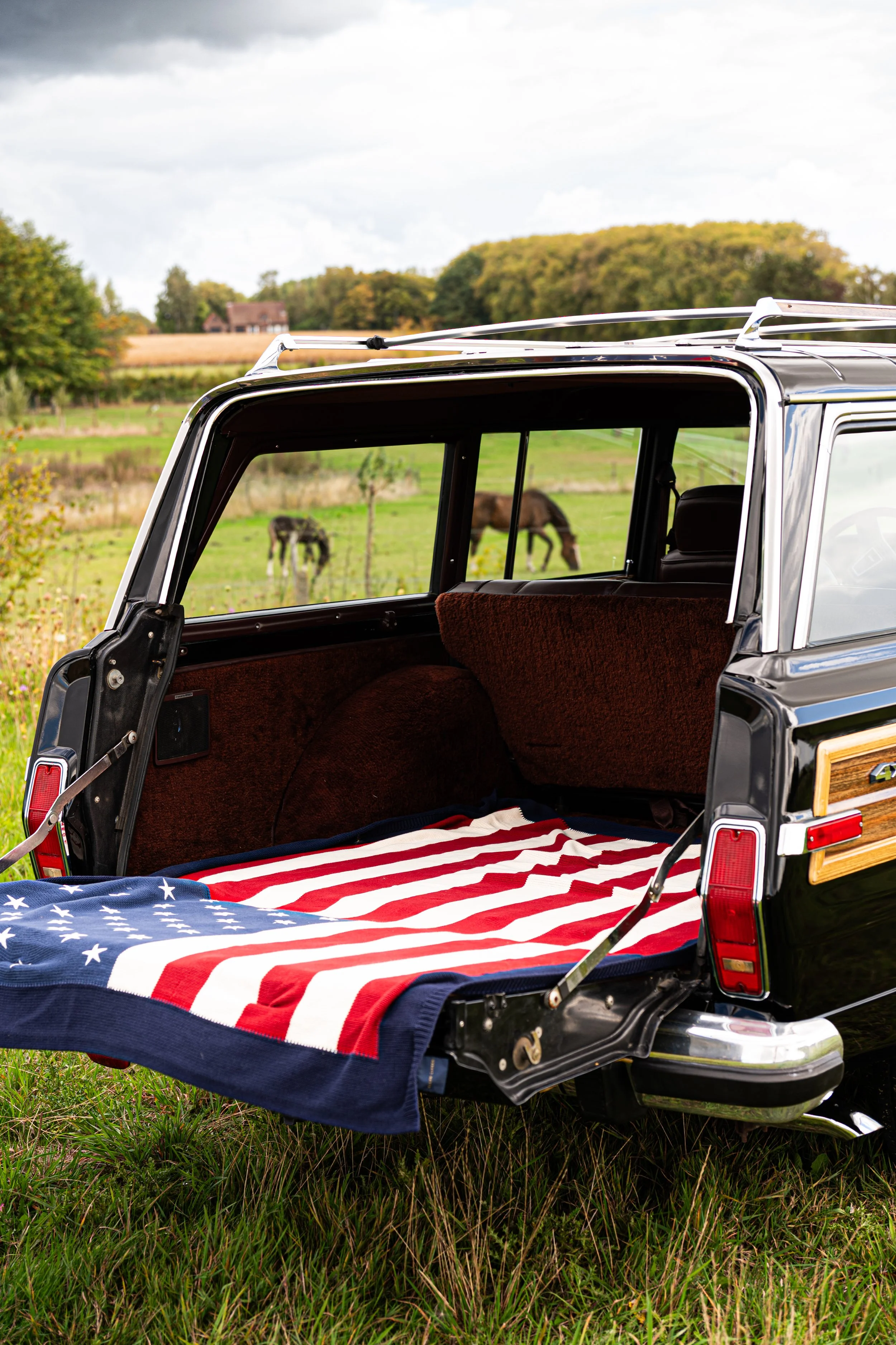 Open rear of a black vintage station wagon with an American flag blanket inside, parked on grass in a rural field with horses grazing in the background.