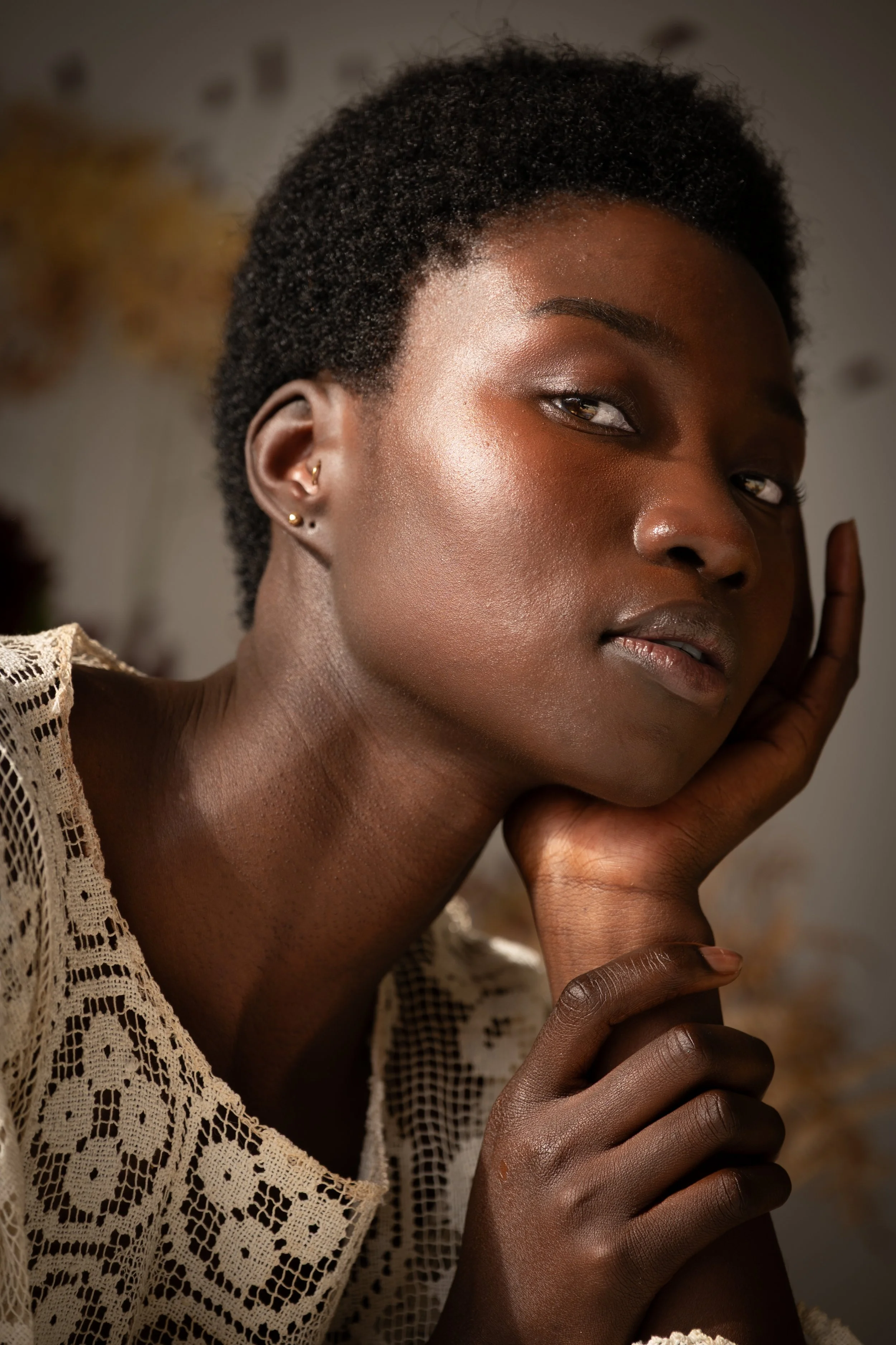 Close-up portrait of a woman with short, curly hair, wearing earrings, resting her face on her hand, wearing a cream lace top.