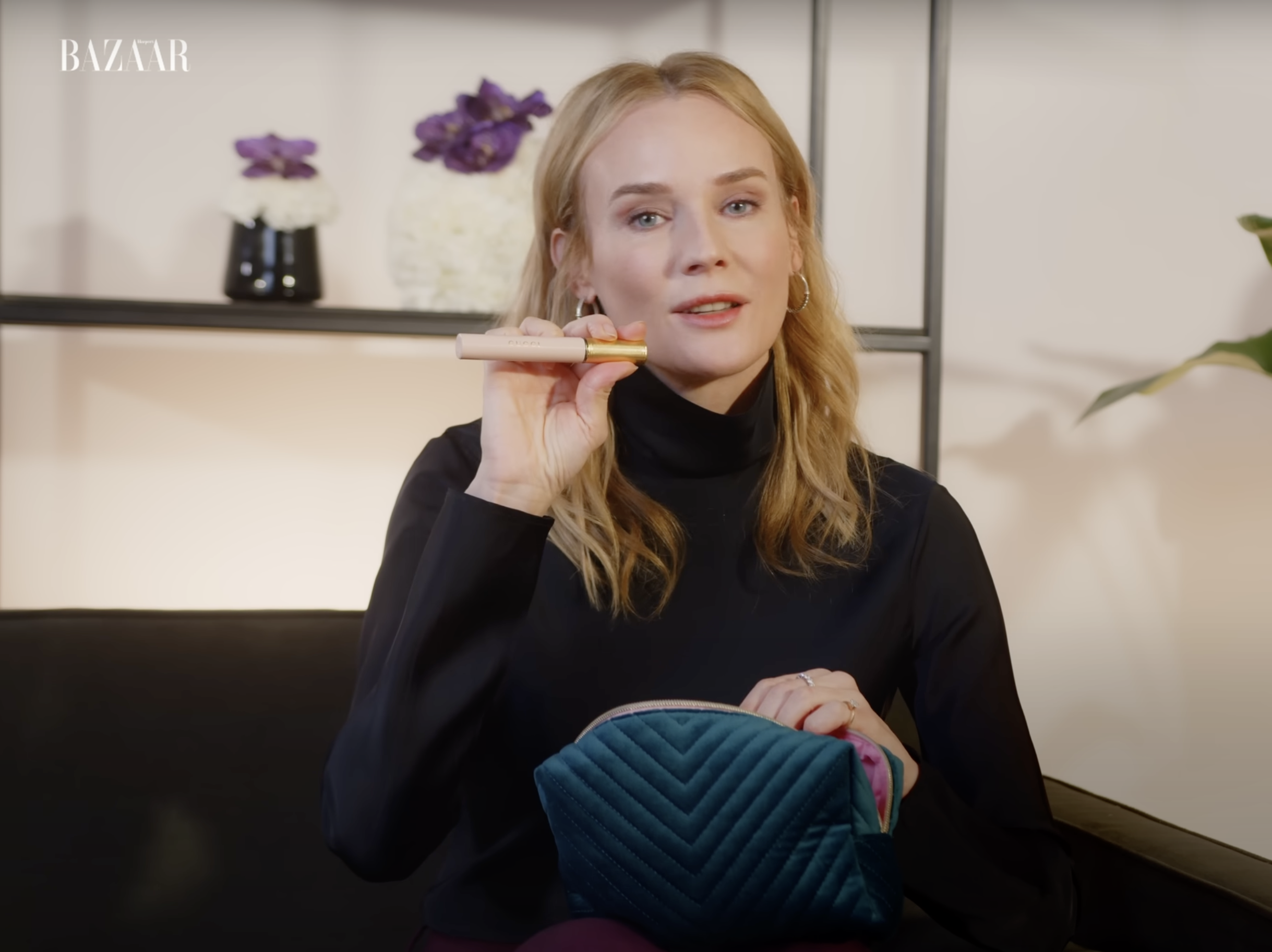 A woman with blonde hair wearing a black turtleneck sitting on a black couch, holding a pink and gold bottle to her face, with a blue quilted bag on her lap, in a room with flowers on shelves in the background.