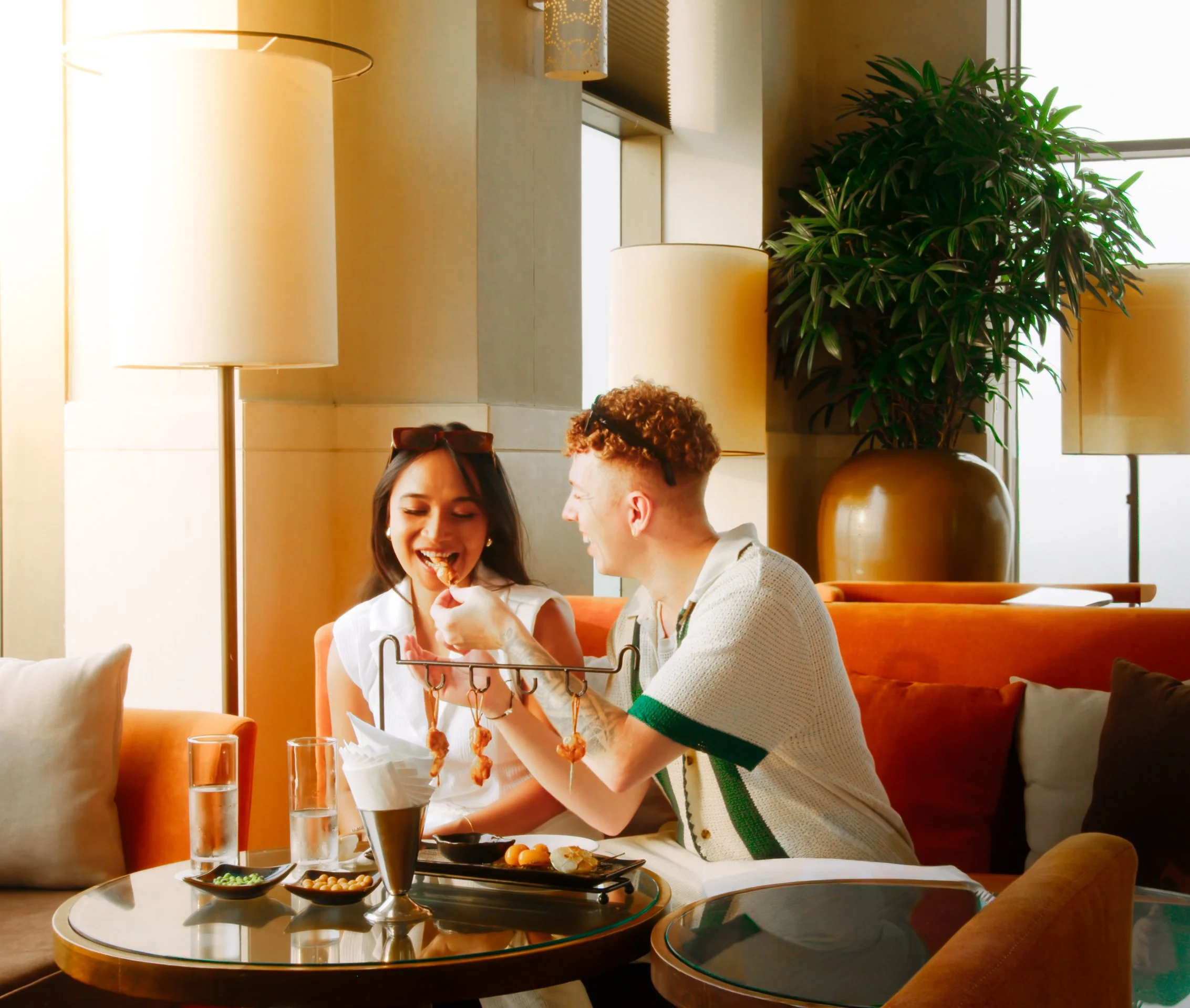 A young man feeds sushi to a woman sitting in a restaurant with a large plant and lamps in the background.