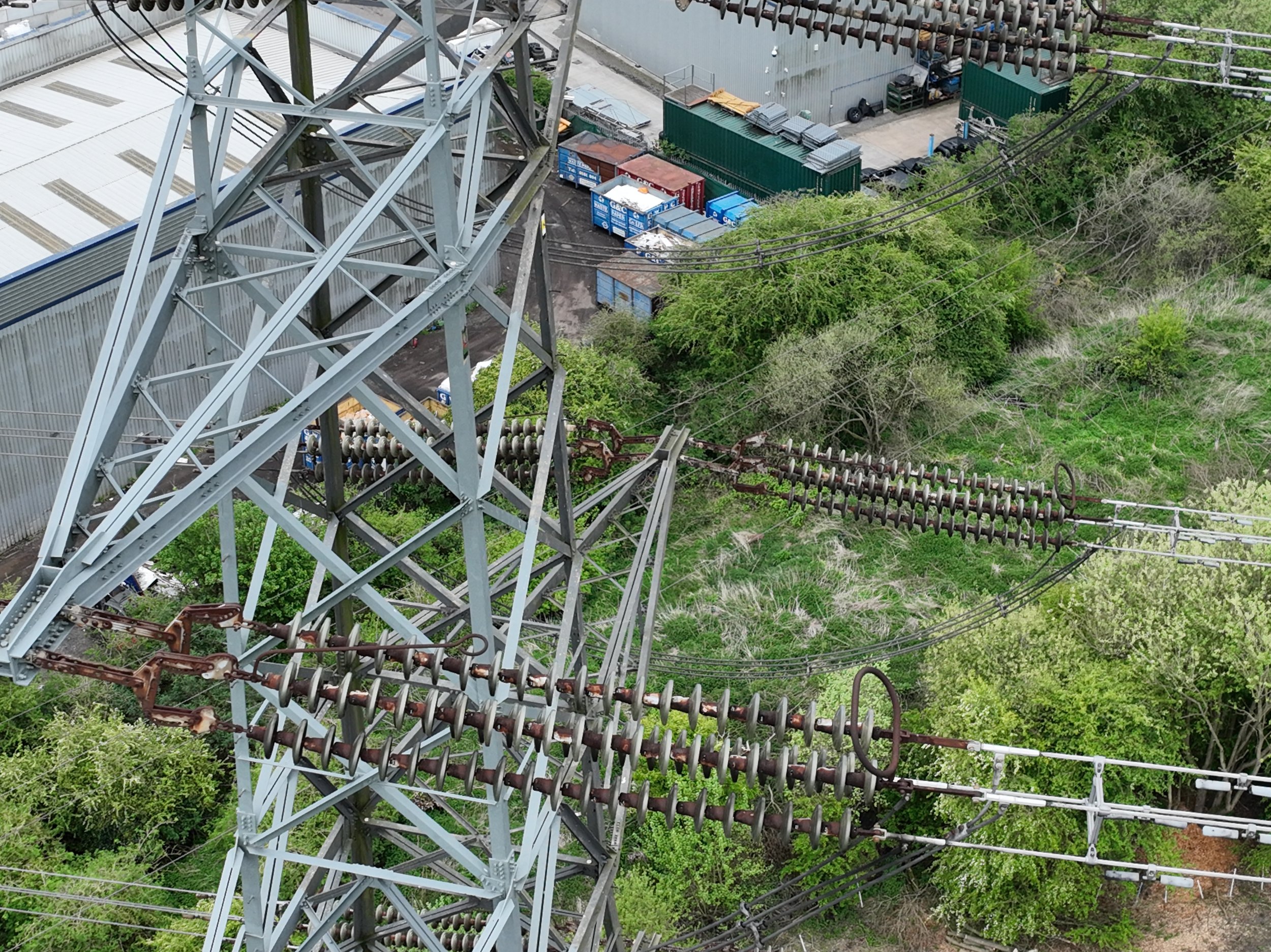 Aerial view of a tall electrical transmission tower with power lines and insulators surrounded by green trees and industrial buildings in the background.