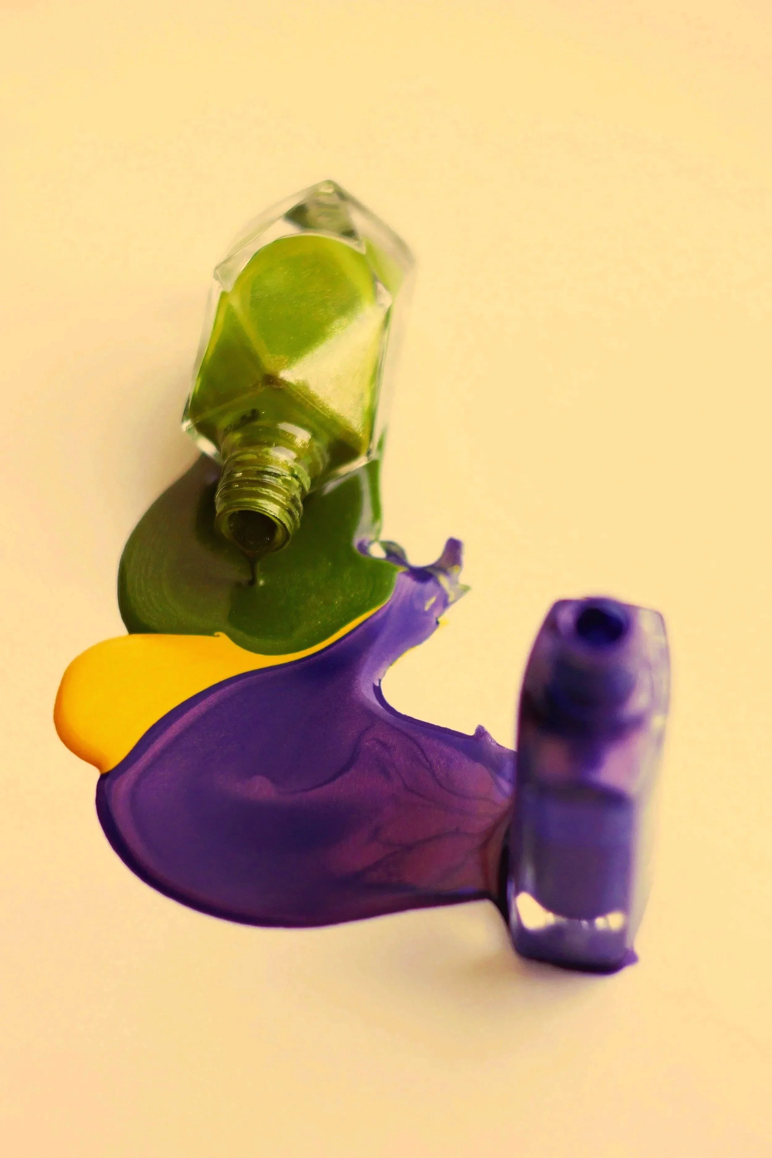 Colorful spilled paint with green, yellow, purple, and blue on a white surface, with a green glass bottle with a drop of paint at the opening.