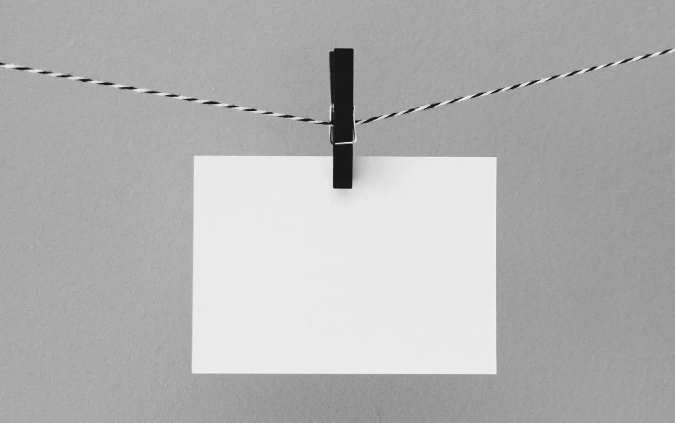A blank white piece of paper hanging from a black clothespin on a string against a gray background.