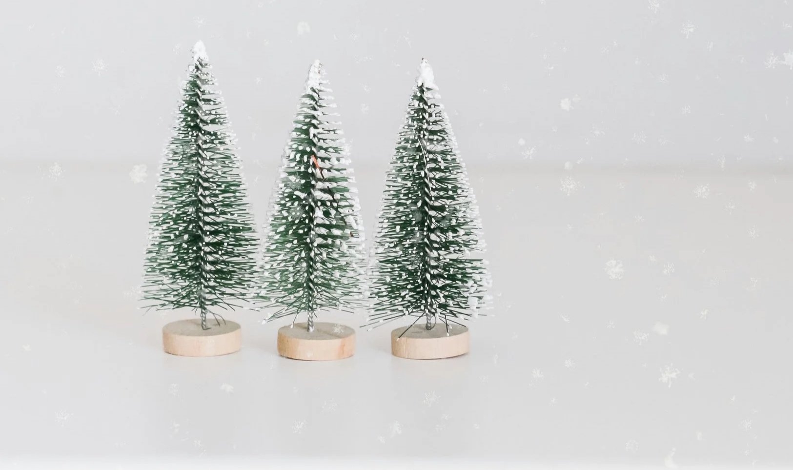 Three small artificial Christmas trees with snow on top, standing on round wooden bases, against a blurry white background with snowfall effects.