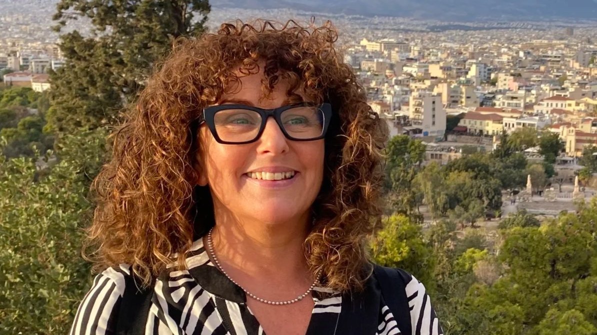 Smiling woman with curly red hair and glasses outdoors with cityscape and trees in the background.
