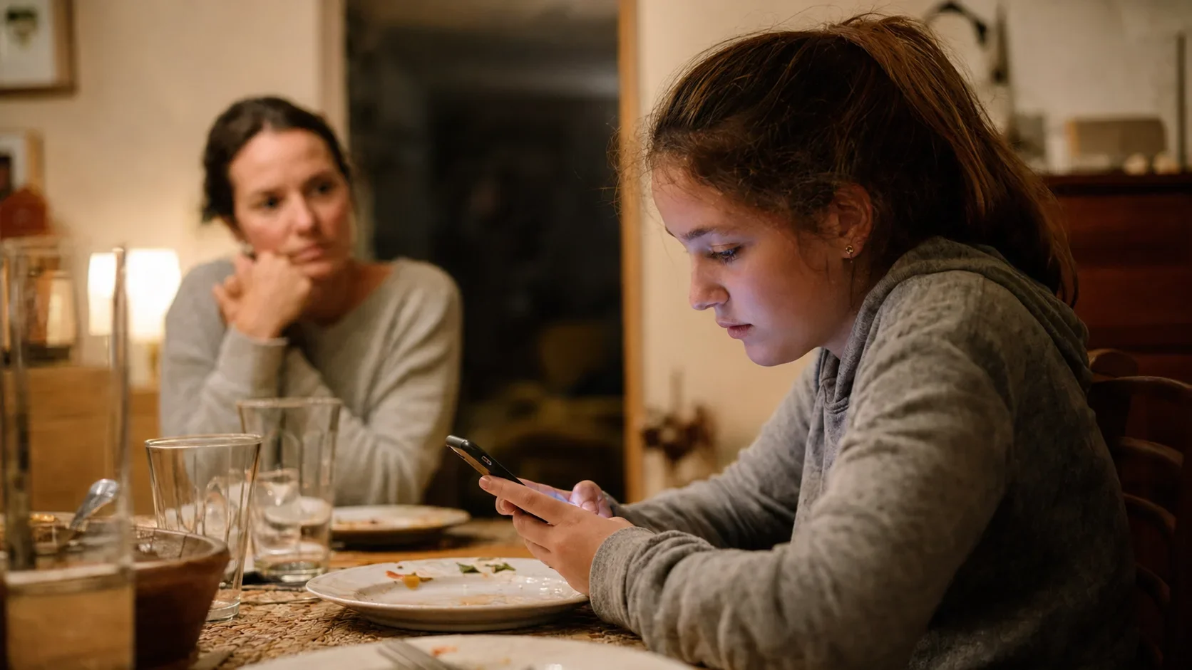 Jugendliche sitzt nach dem Abendessen am Familientisch und schaut konzentriert auf ihr Smartphone, während eine Mutter im Hintergrund besorgt zu ihr blickt.