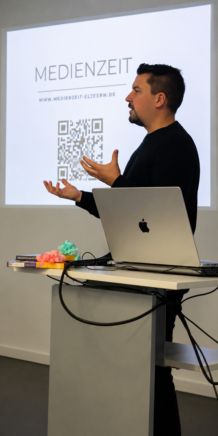 Tobias Dillinger bei einem Medienzeit Vortrag in einem modernen Seminarraum. Er steht neben einem Laptop an einem Rednerpult und spricht gestikulierend vor einer projizierten Medienzeit Präsentation mit QR-Code im Hintergrund.