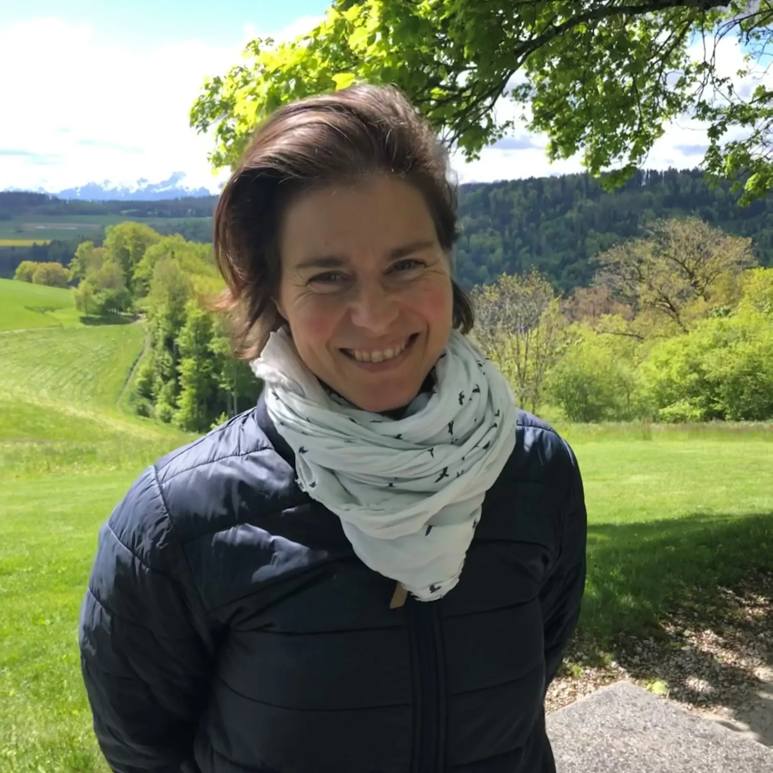Alexandra Berchtold mit dunkler Jacke und hellem Schal steht unter einem Baum. Im Hintergrund liegen grüne Wiesen, bewaldete Hügel und Berge