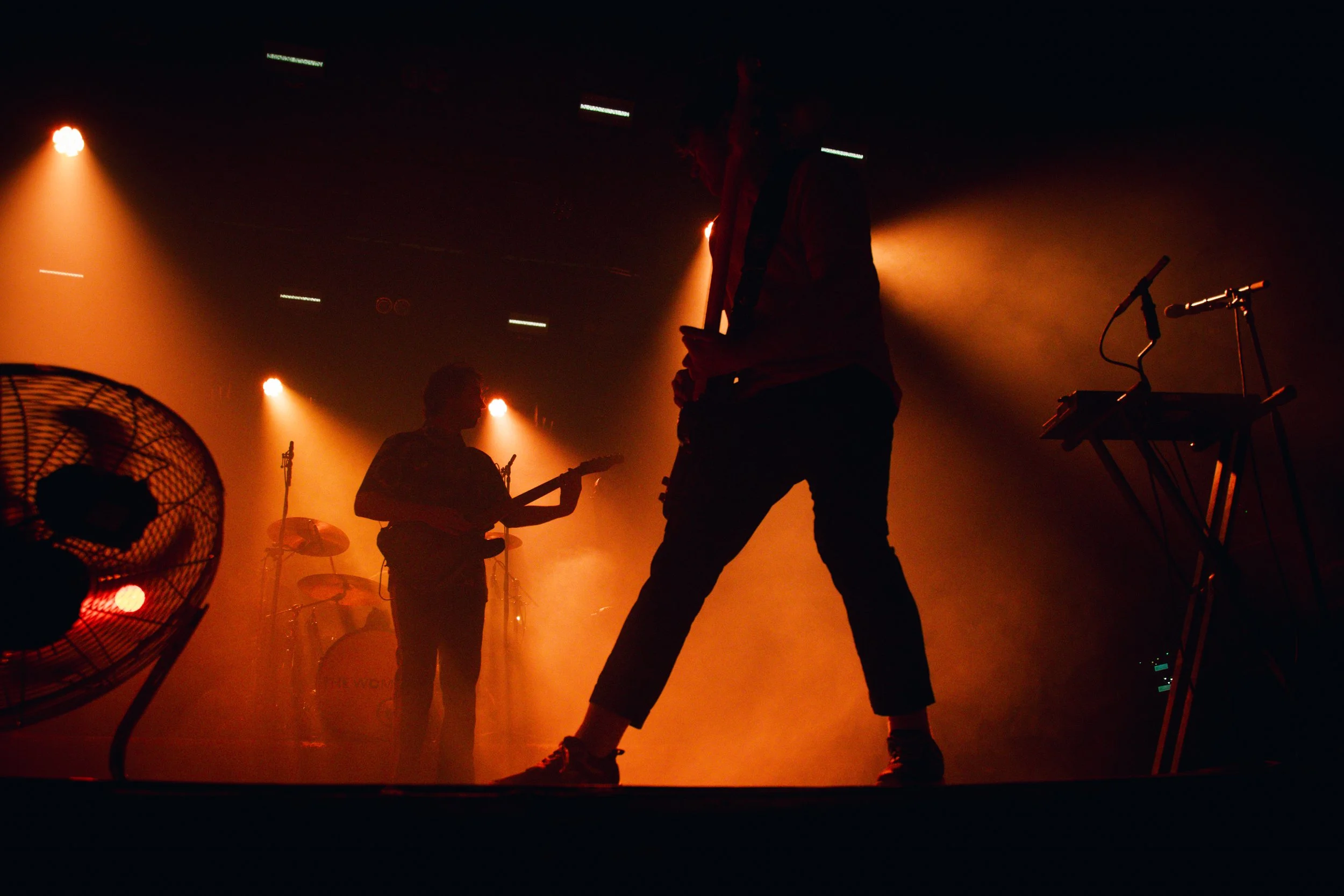 Silhouettes of musicians playing on stage with orange and yellow lighting, a fan, and musical equipment in the background.