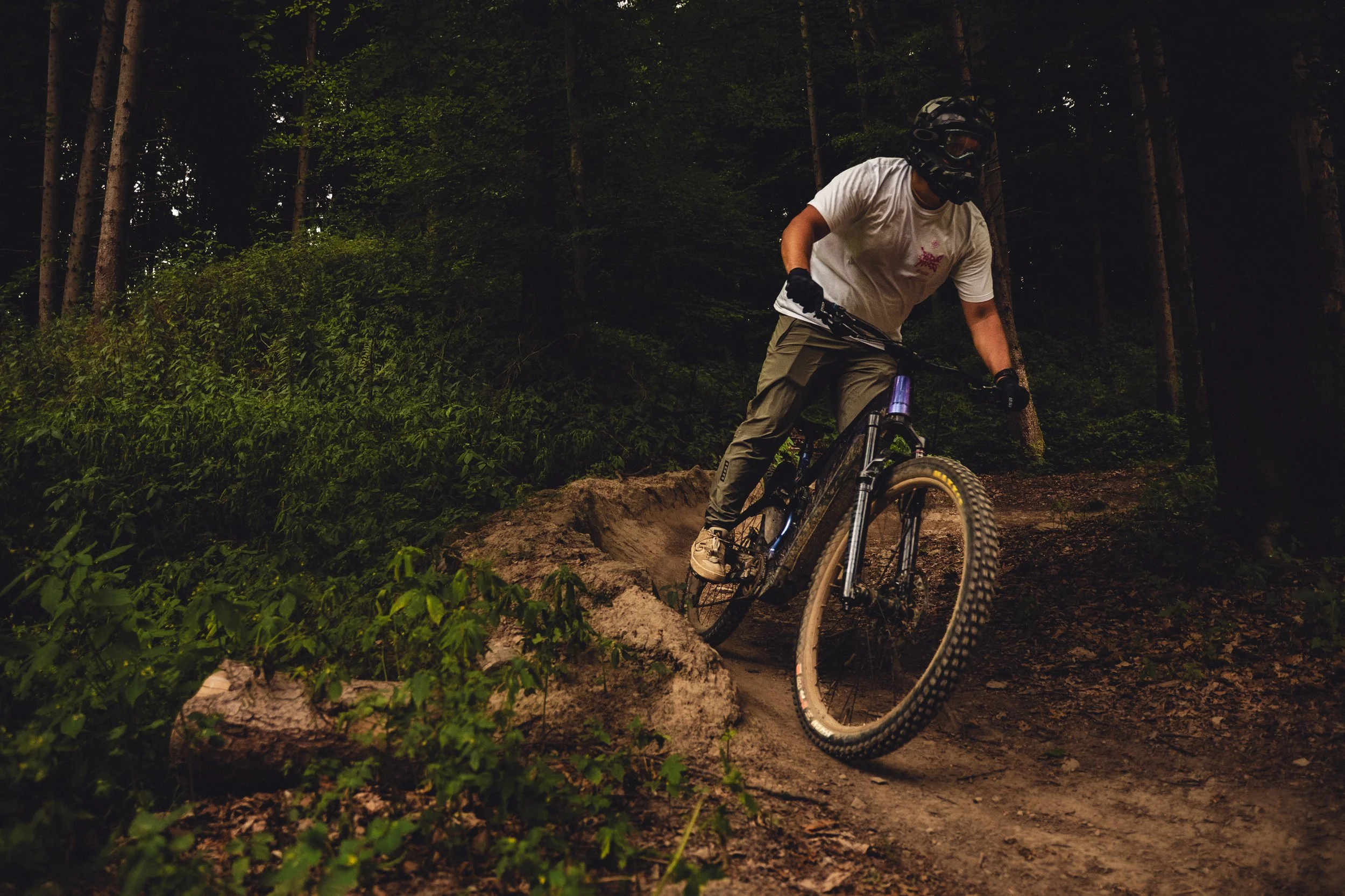 A mountain biker wearing a helmet and white t-shirt riding on a dirt trail in a dense forest.