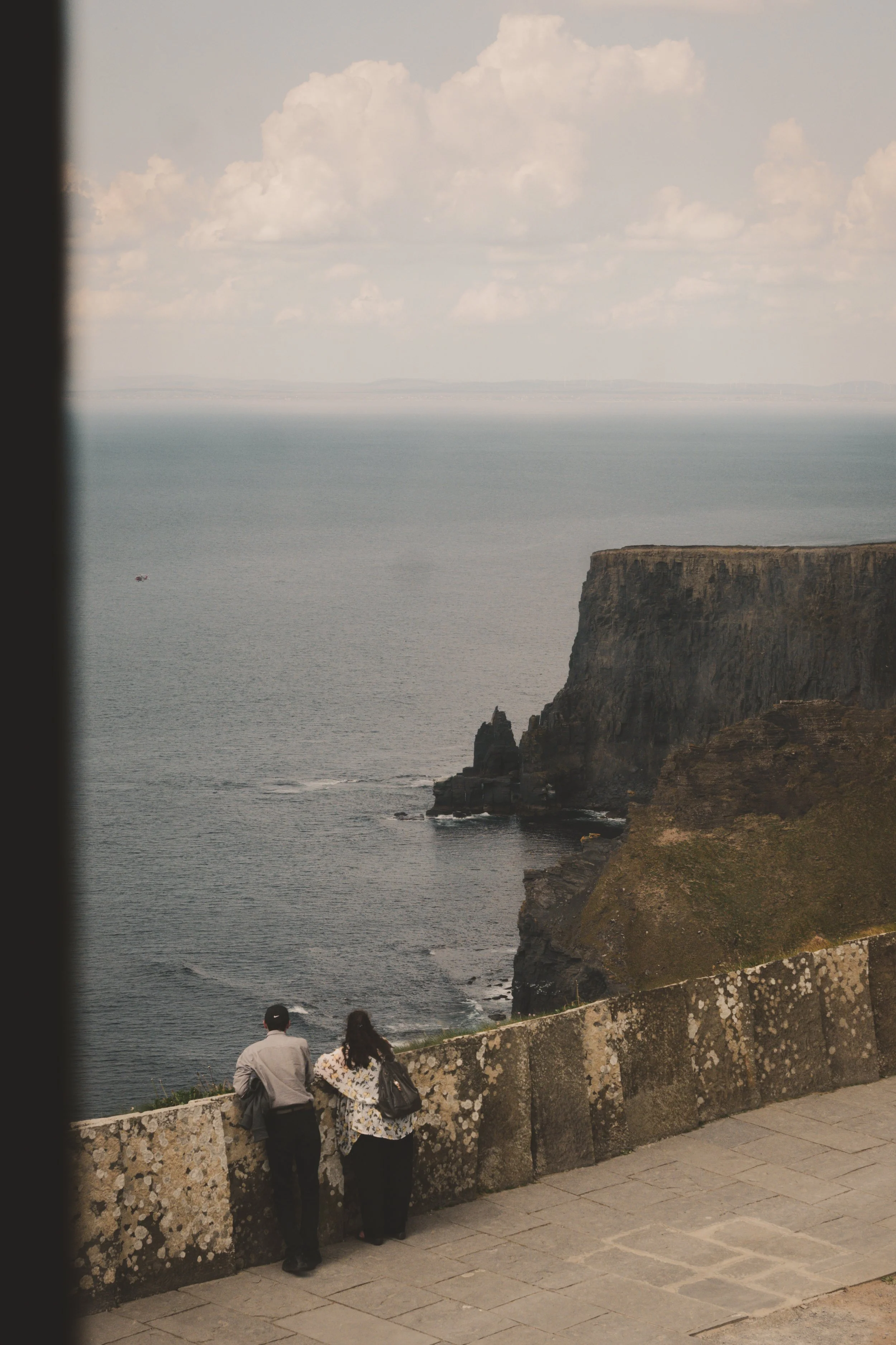 Two people, a man and a woman, standing by a stone railing overlooking the ocean with cliffs in the background.