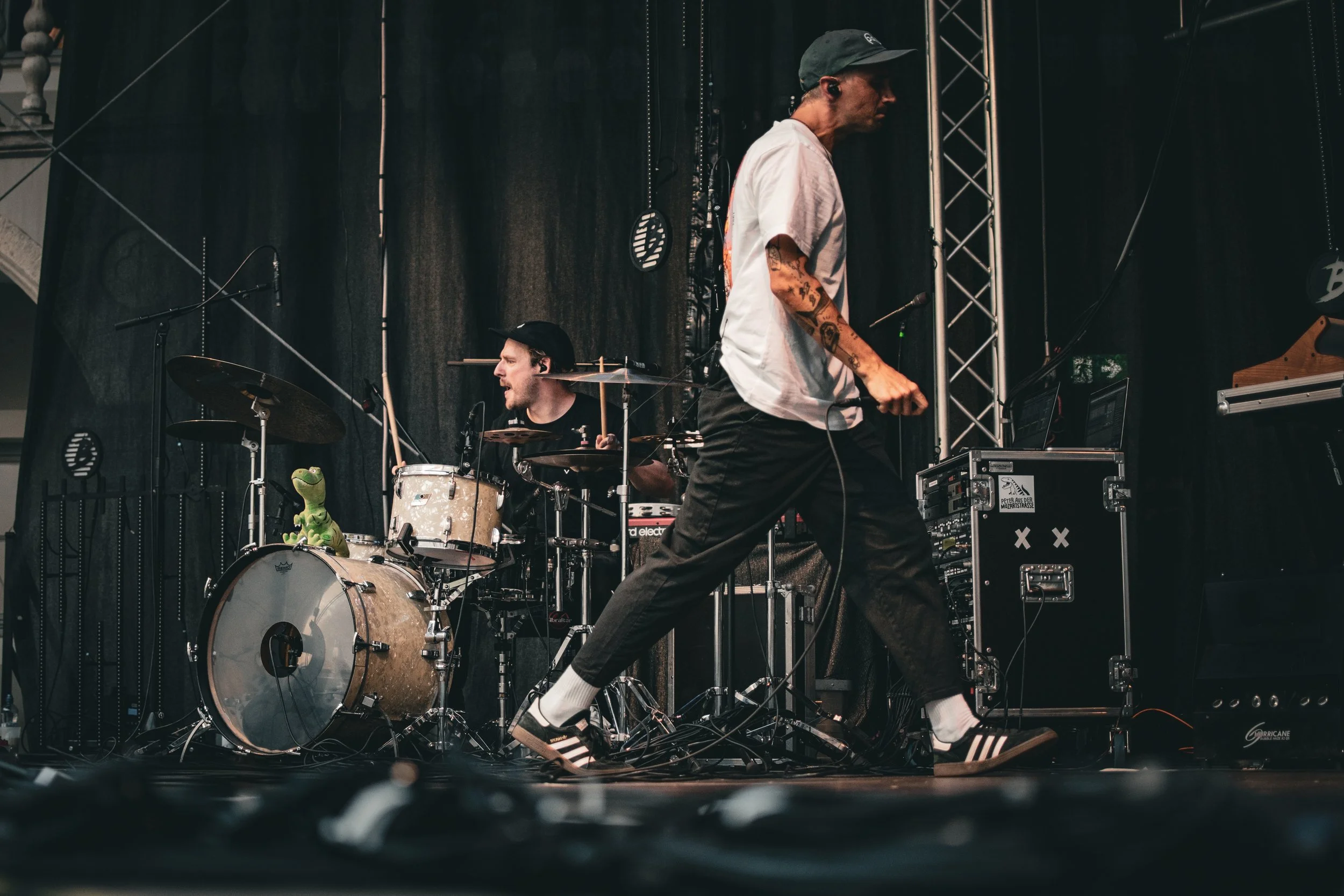 Two musicians performing on stage. One is a drummer behind a drum set with a green dinosaur toy on the bass drum. The other is a standing performer with tattoos, wearing a white t-shirt, black pants, white socks, and Adidas sneakers, holding a microp