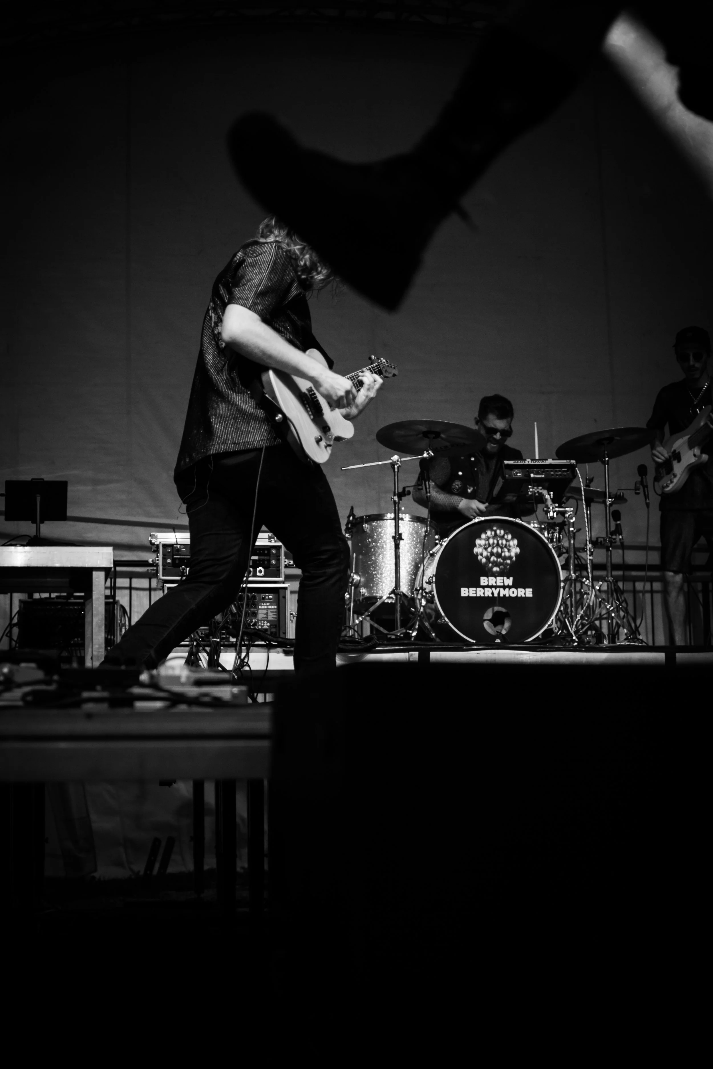 A black and white photo of a band performing on stage, with a guitarist in the foreground, a drummer in the middle, and a bassist in the background. The drum kit has a logo that reads 'Brew Berrymore'.