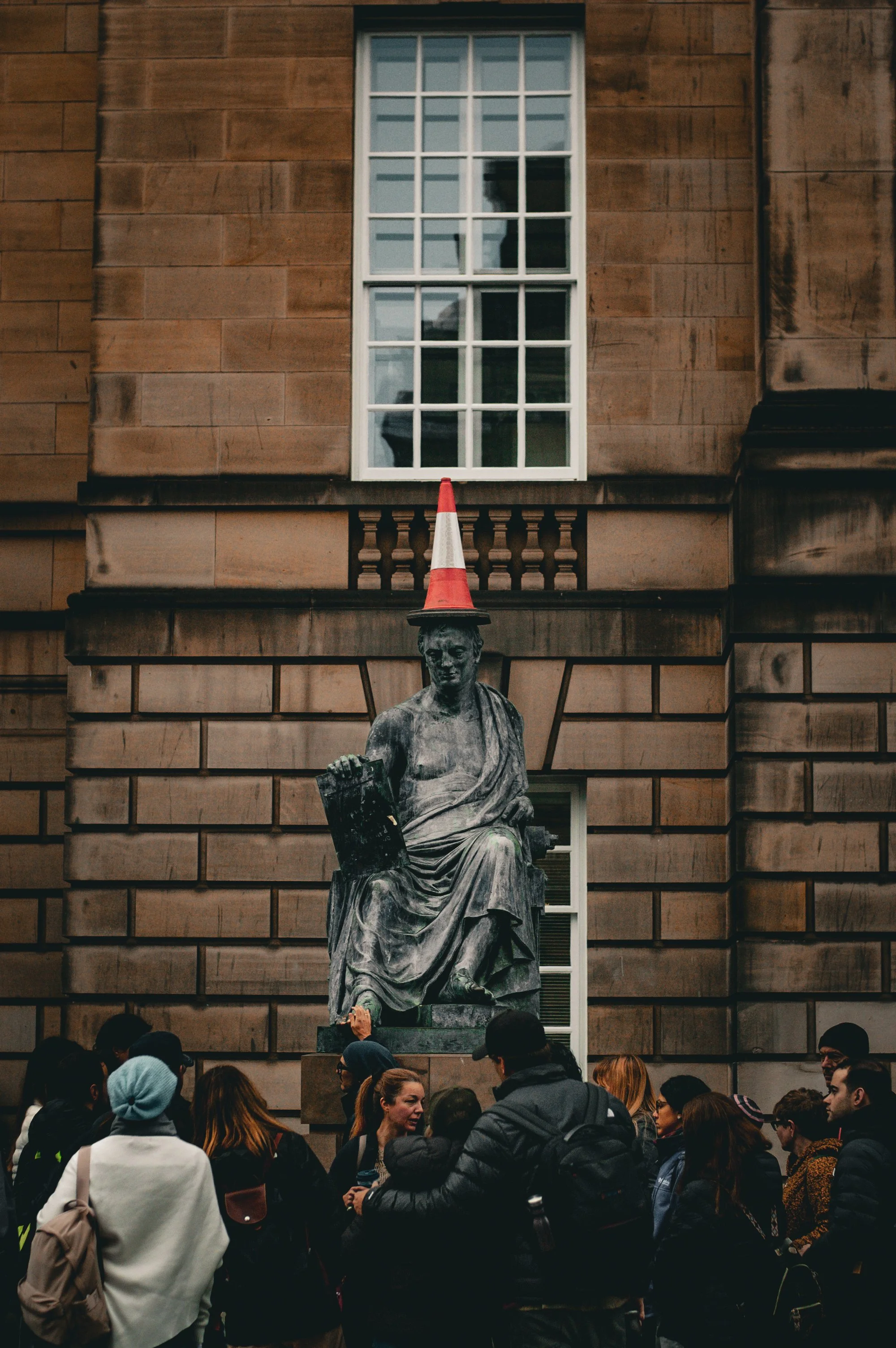 A stone statue of a seated woman with a book, topped with a traffic cone, standing in front of a large architecture building with a window, and surrounded by a crowd of people.