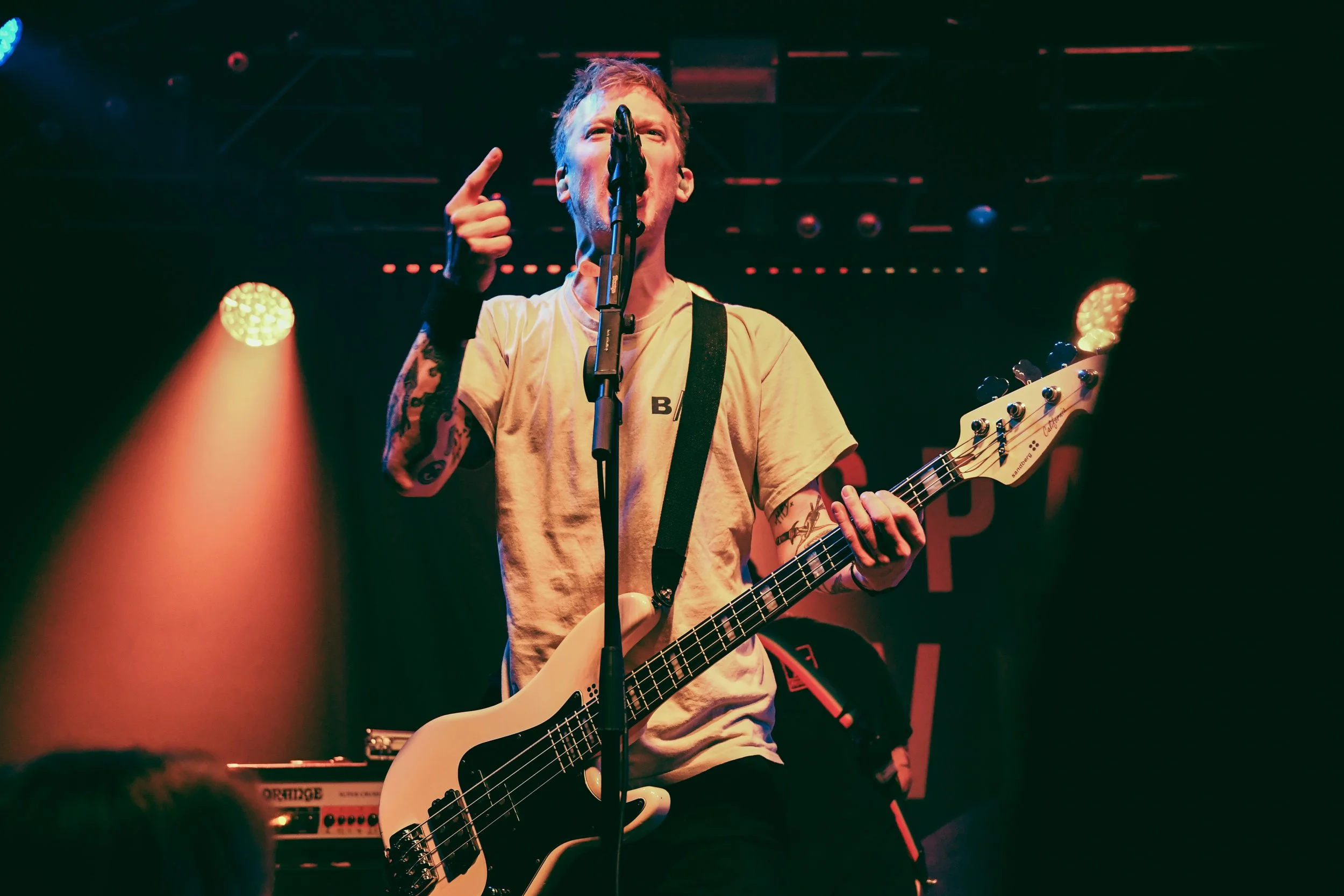 A musician with tattoos plays a bass guitar on stage, singing into a microphone, with stage lights illuminating the scene.