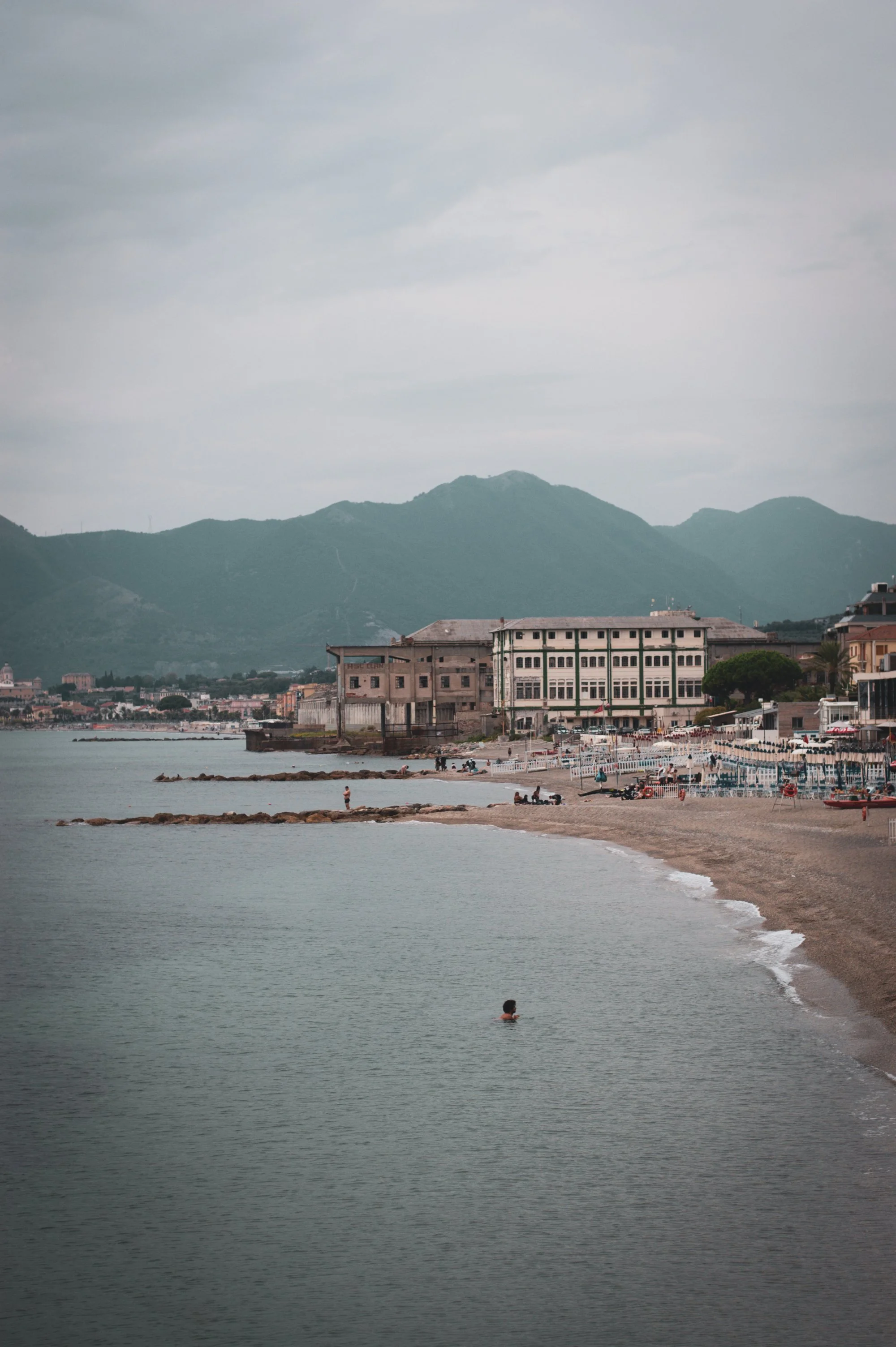 A person swimming in the calm water near a sandy beach with buildings and mountains in the background.