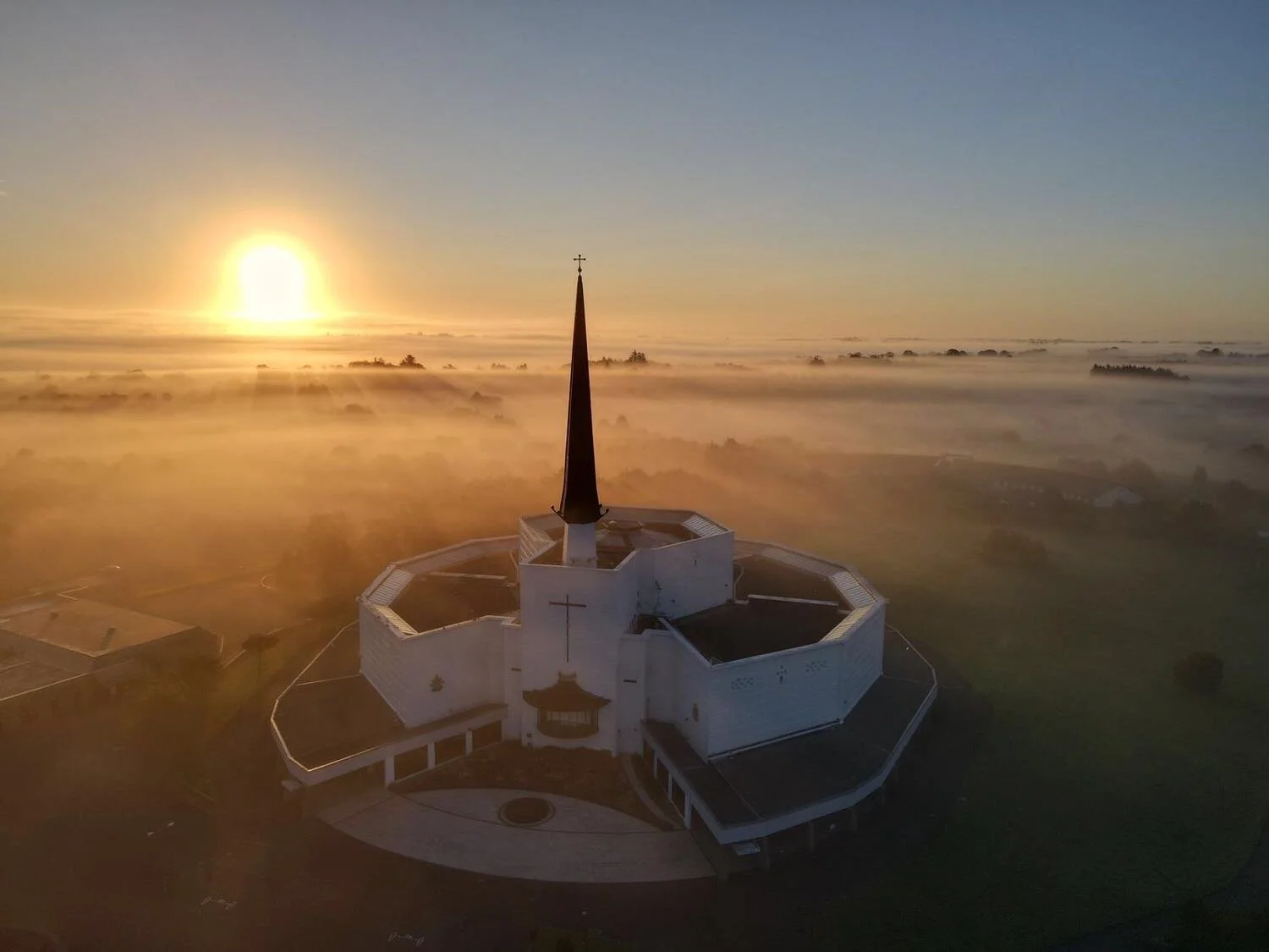 A church with a tall steeple and a cross at sunset or sunrise, surrounded by fog over a landscape with scattered houses and trees.