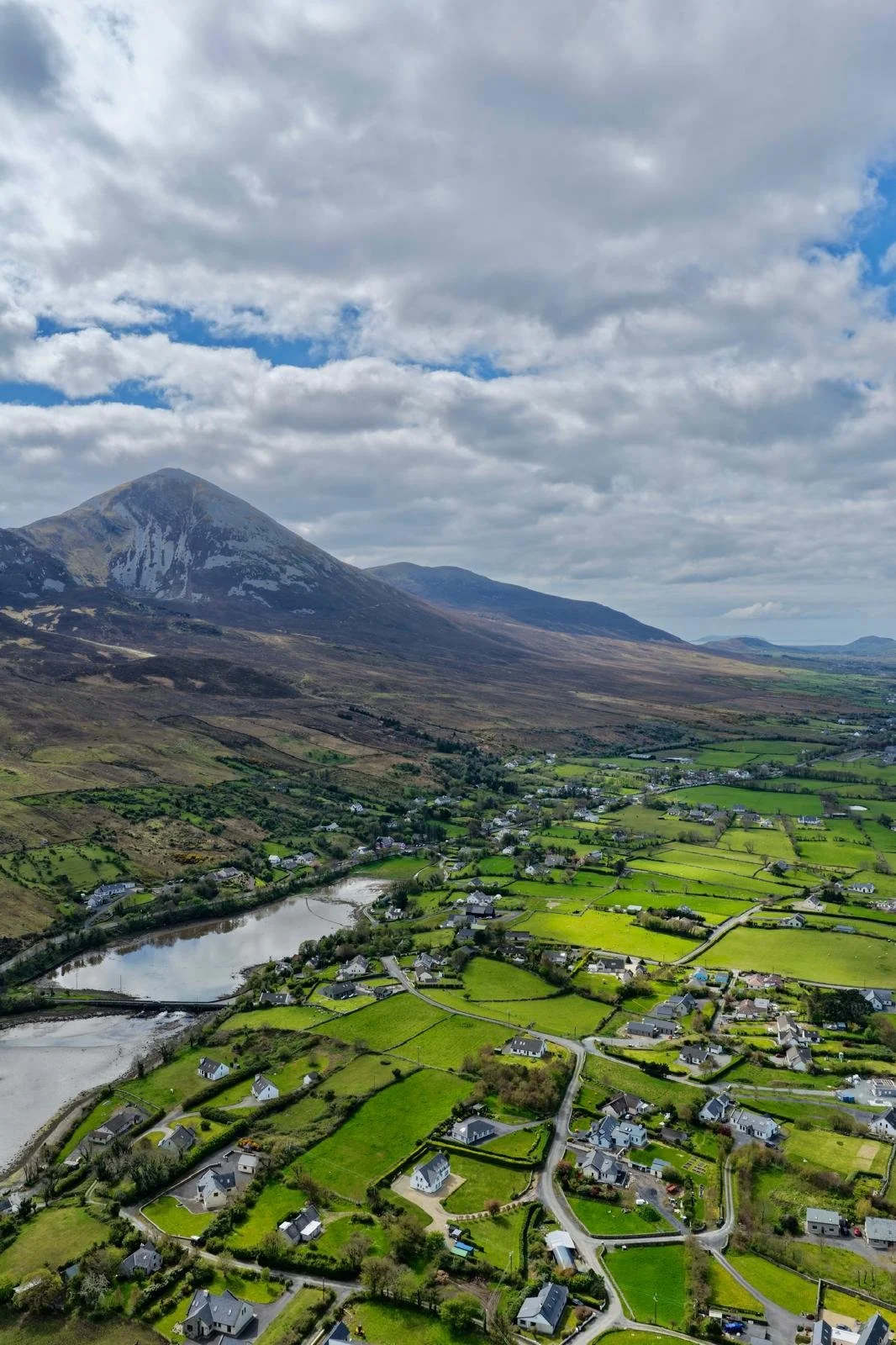 Aerial view of a green valley with scattered houses, a river, and a large mountain in the background under a partly cloudy sky.