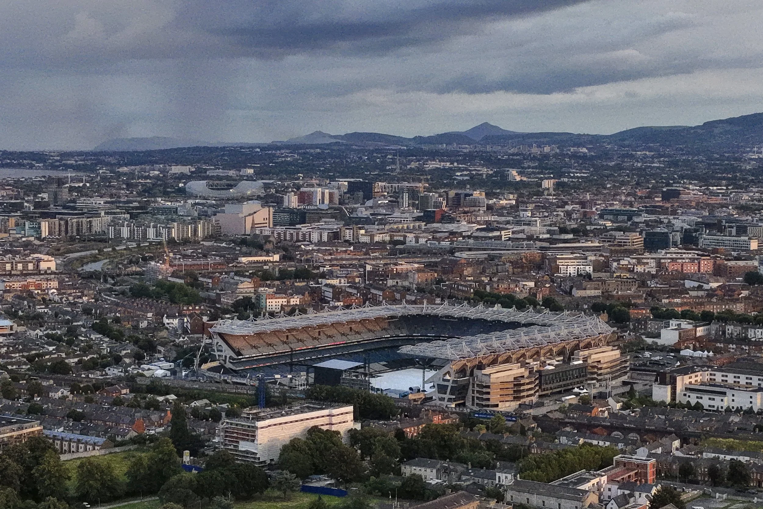 A panoramic view of a city with a large stadium in the foreground and a dense urban area with residential and commercial buildings. Mountains are visible in the distance under a cloudy sky.