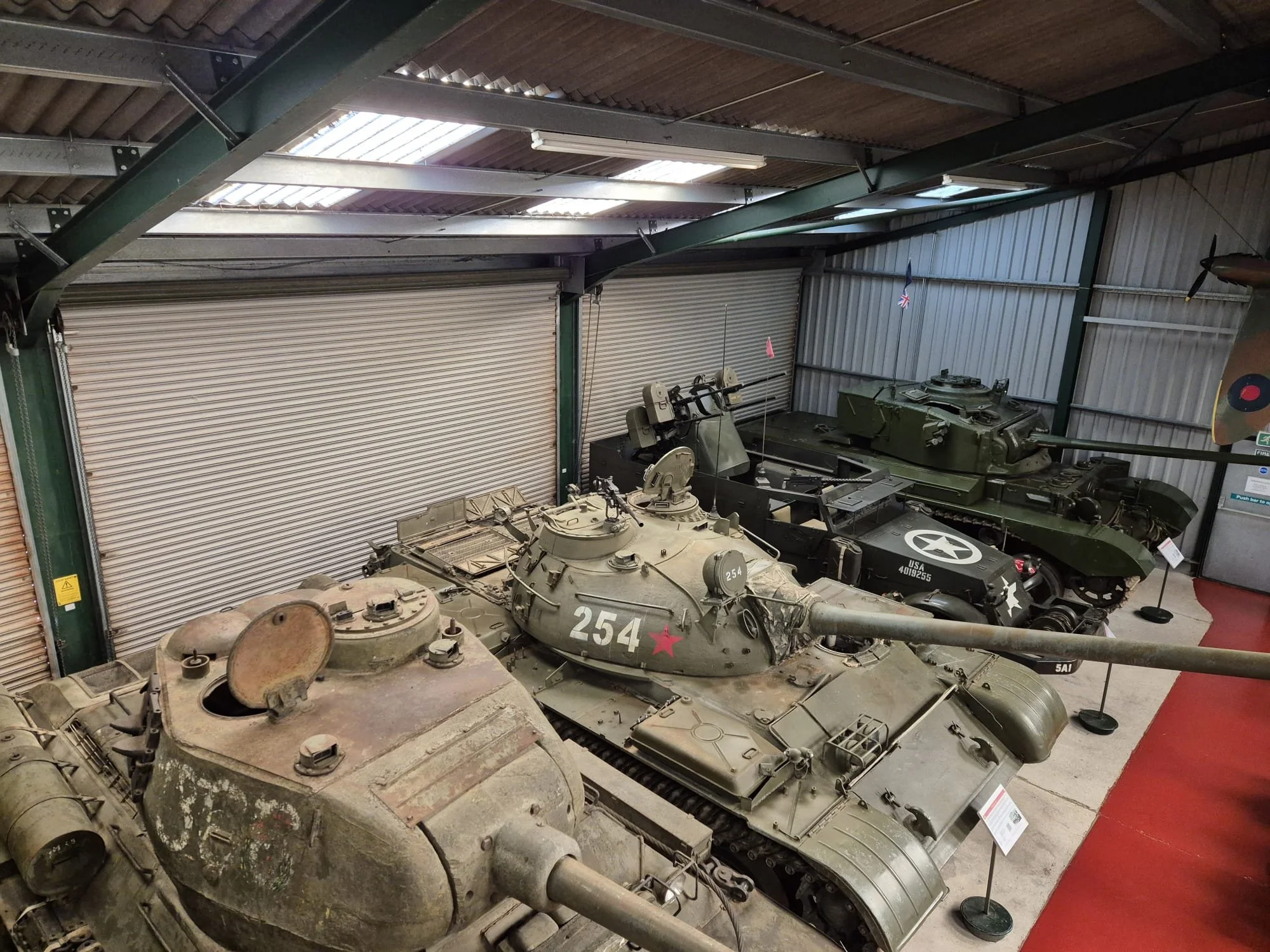 Several military tanks inside a warehouse, including a weathered one in the foreground and others in better condition in the background, with informational placards next to each.