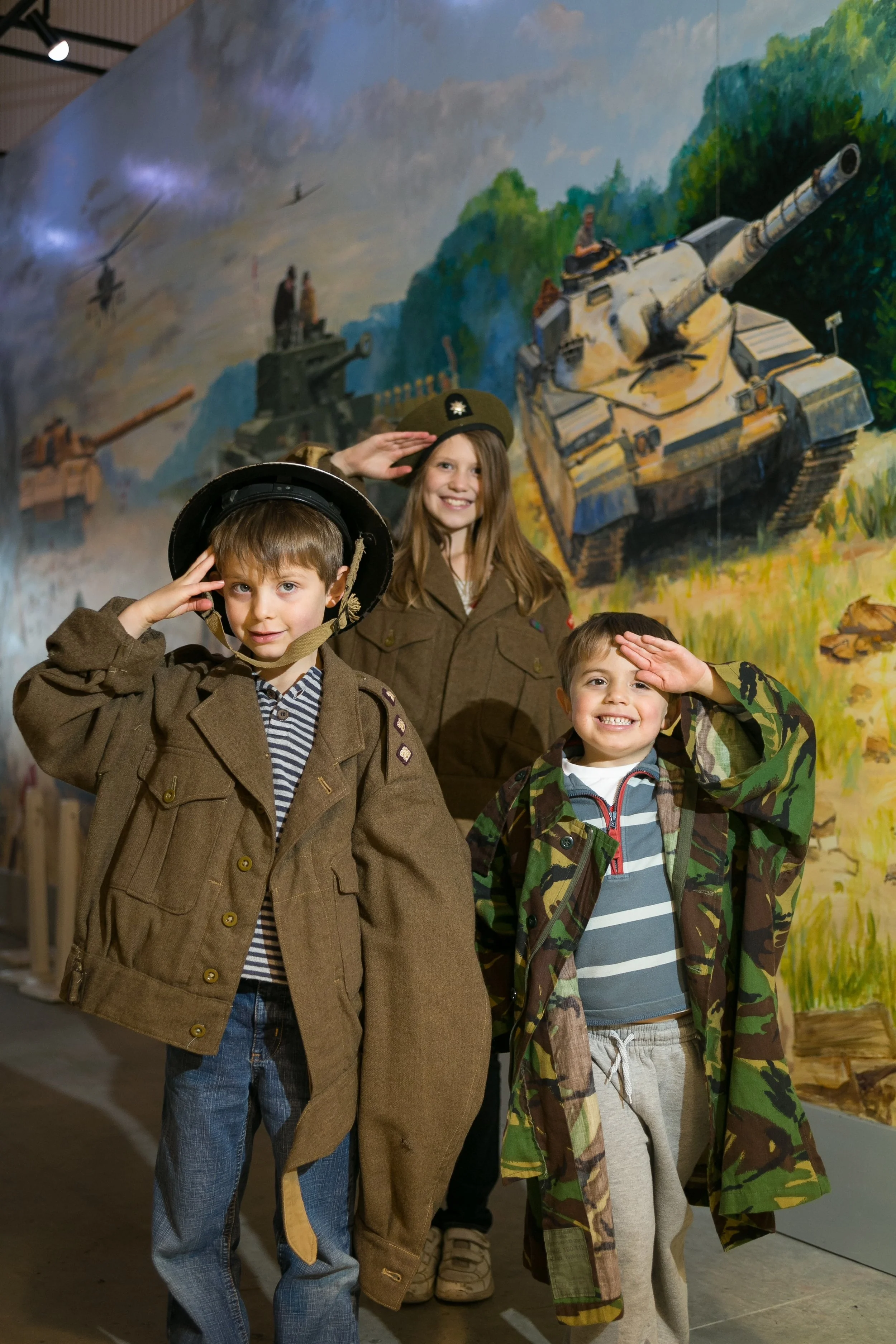 Three children in military costumes saluting in front of a painted World War II battlefield mural.
