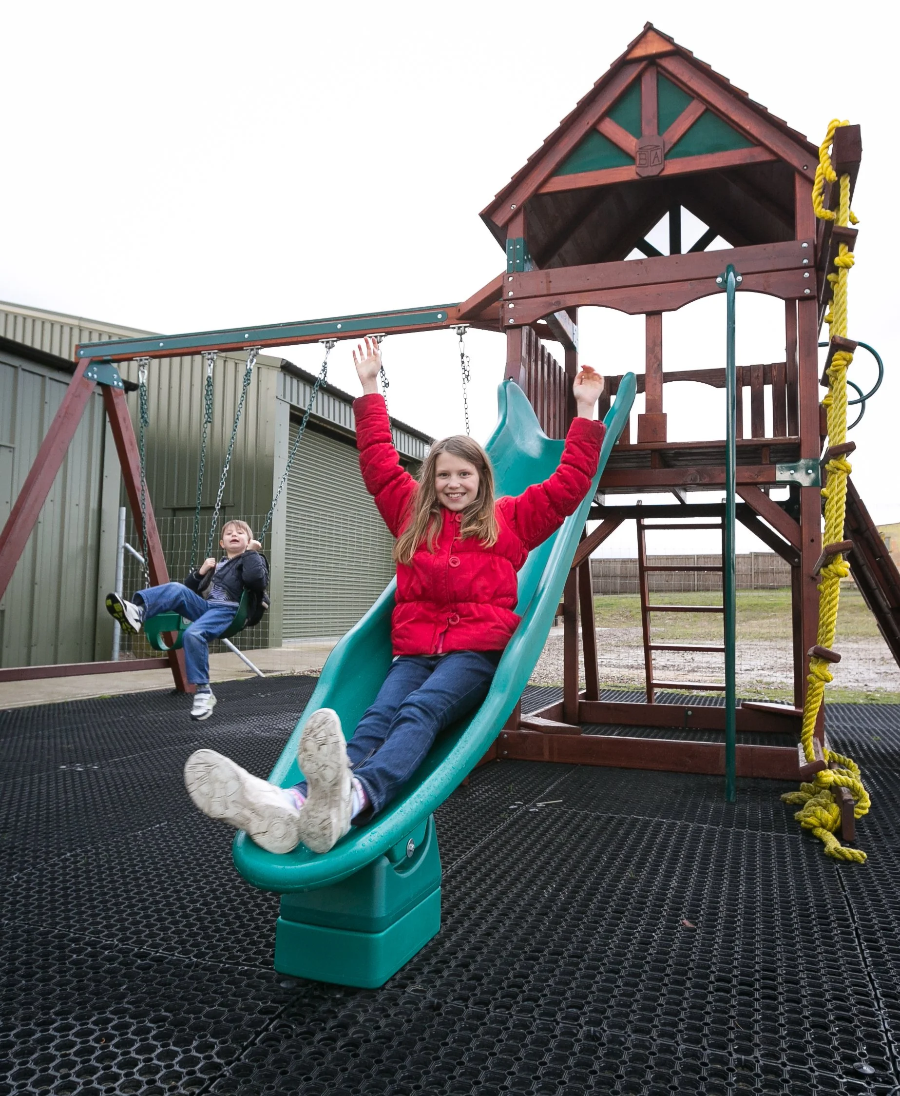Two children playing on a playground with a slide, swings, and climbing structure, outdoors on a cloudy day.