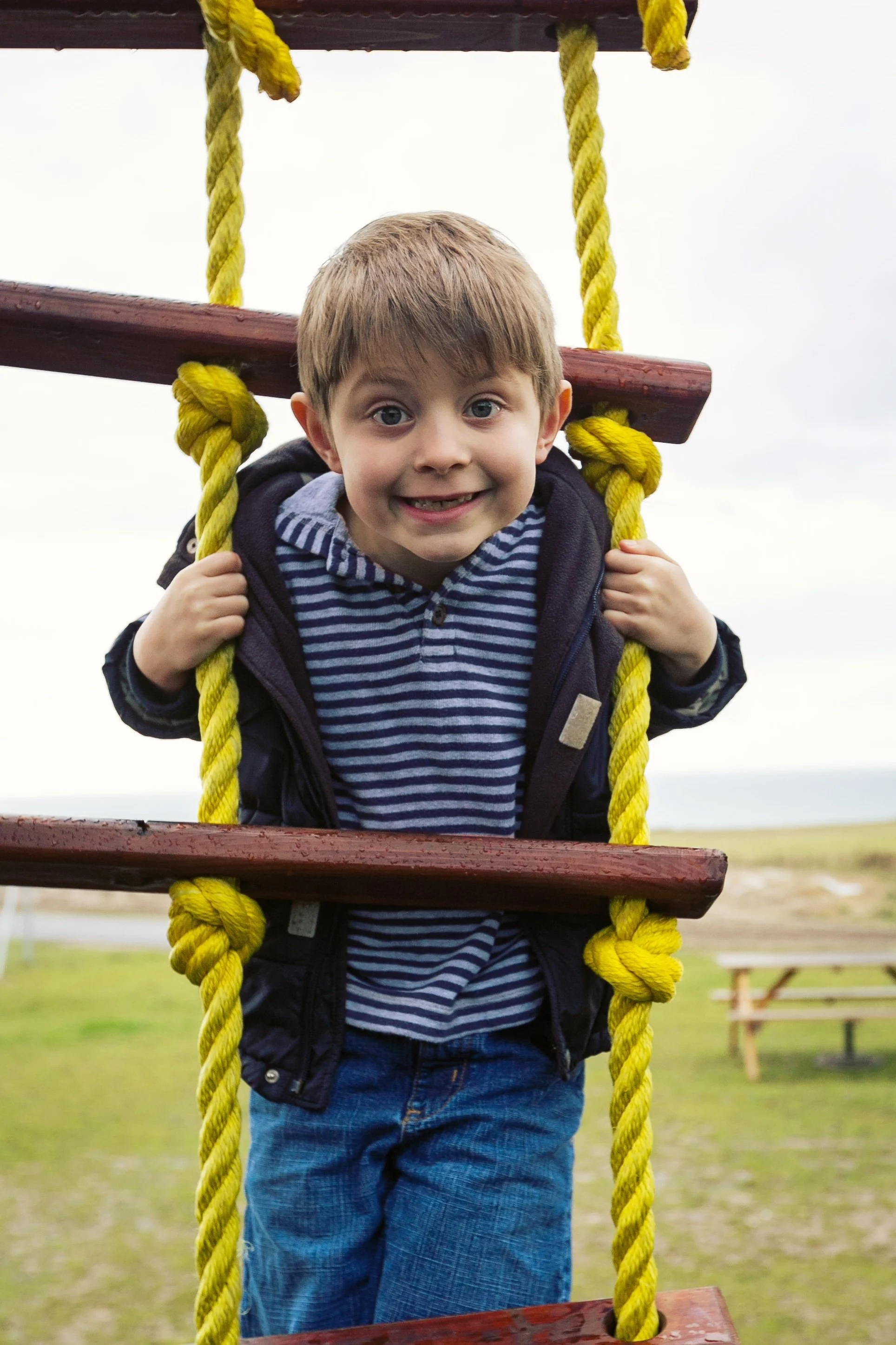 A young boy with light brown hair, smiling, wearing a striped navy and gray hoodie and a dark jacket, climbing a wooden ladder with yellow ropes at an outdoor park.