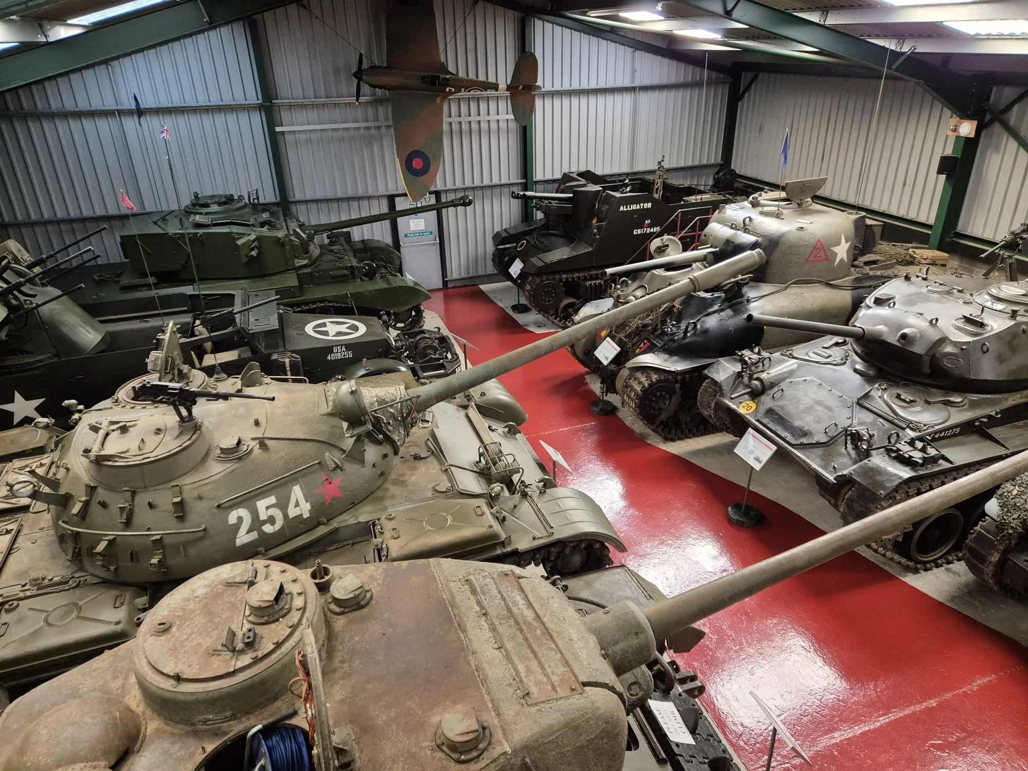 Museum exhibit of various military tanks and a fighter plane inside a hangar, with informational signs in front of each vehicle.