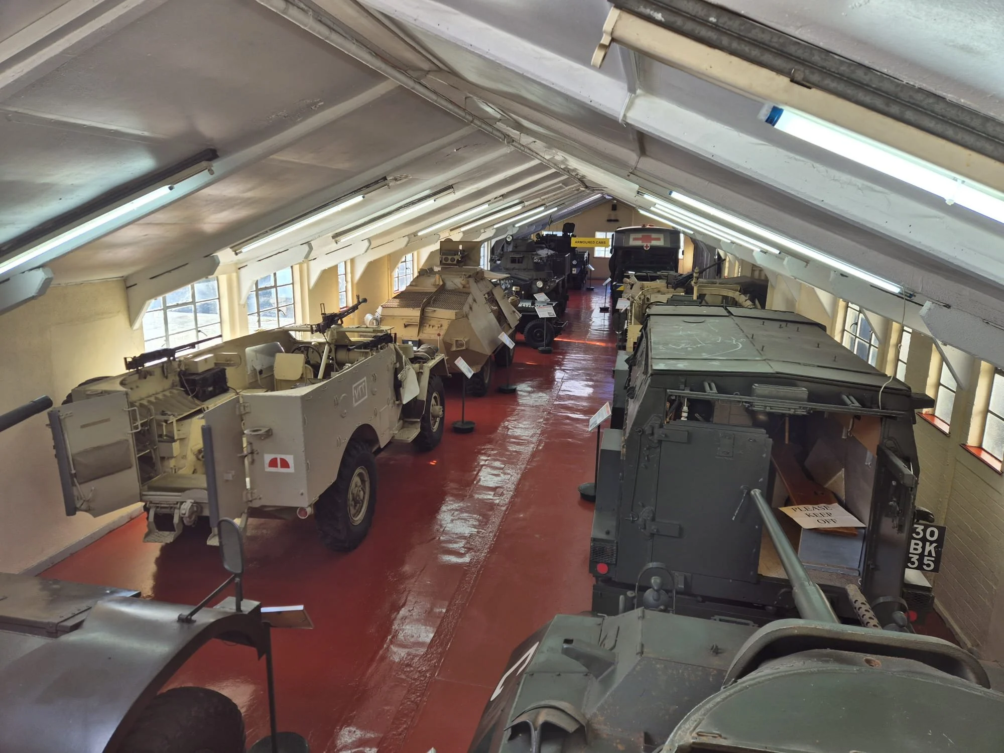 Indoor military vehicle museum display with various armored vehicles arranged in a row under a sloped ceiling with windows, including armored cars, tanks, and military trucks.
