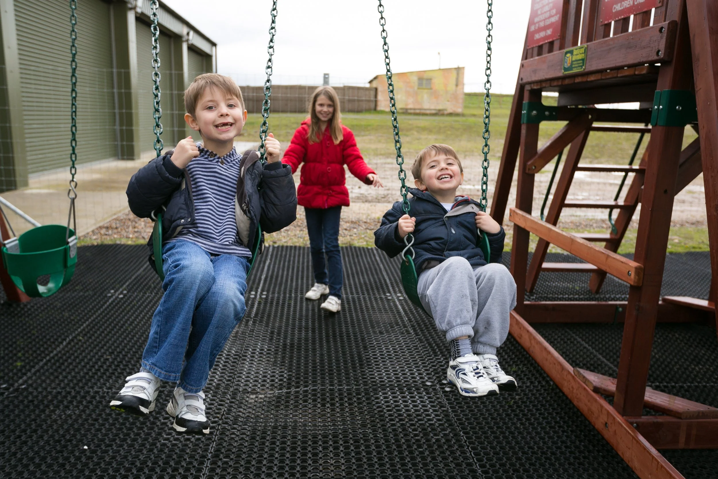 Two young boys on swings and a girl in a red jacket walking behind them at a playground with a wooden slide in the background.