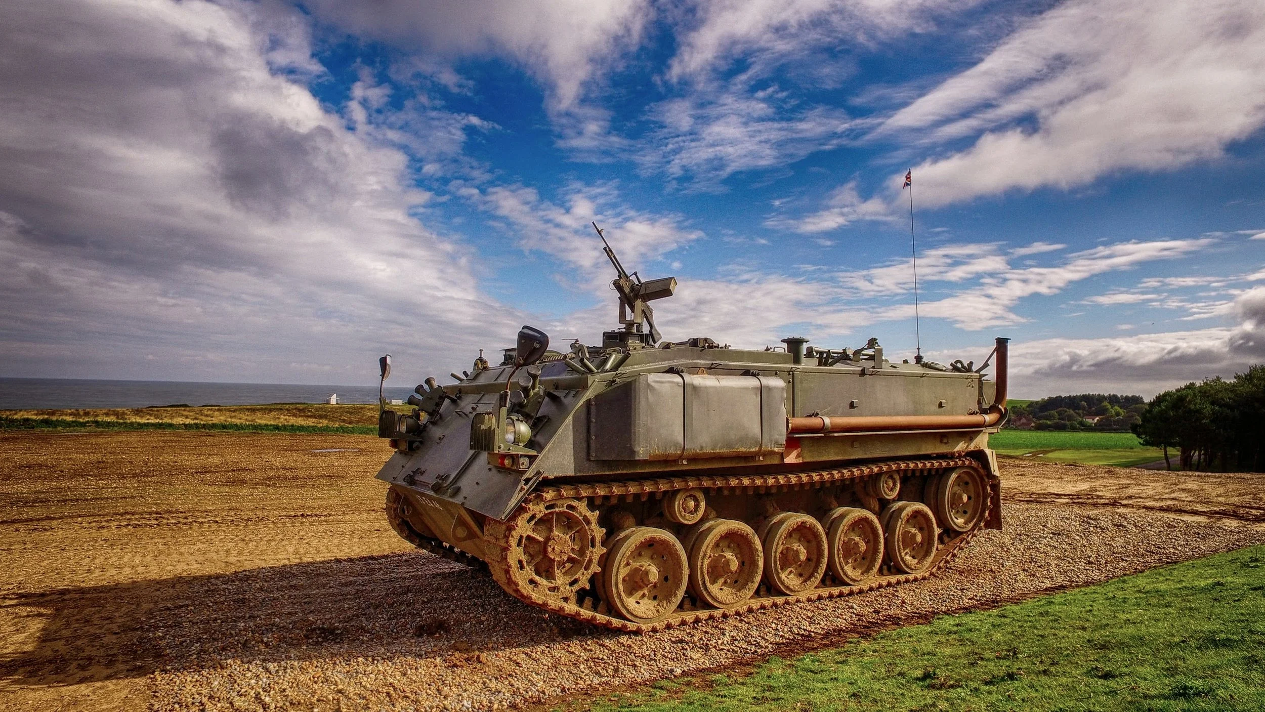 A military tank on a dirt field with a blue sky and scattered clouds, and green grass and trees in the background.
