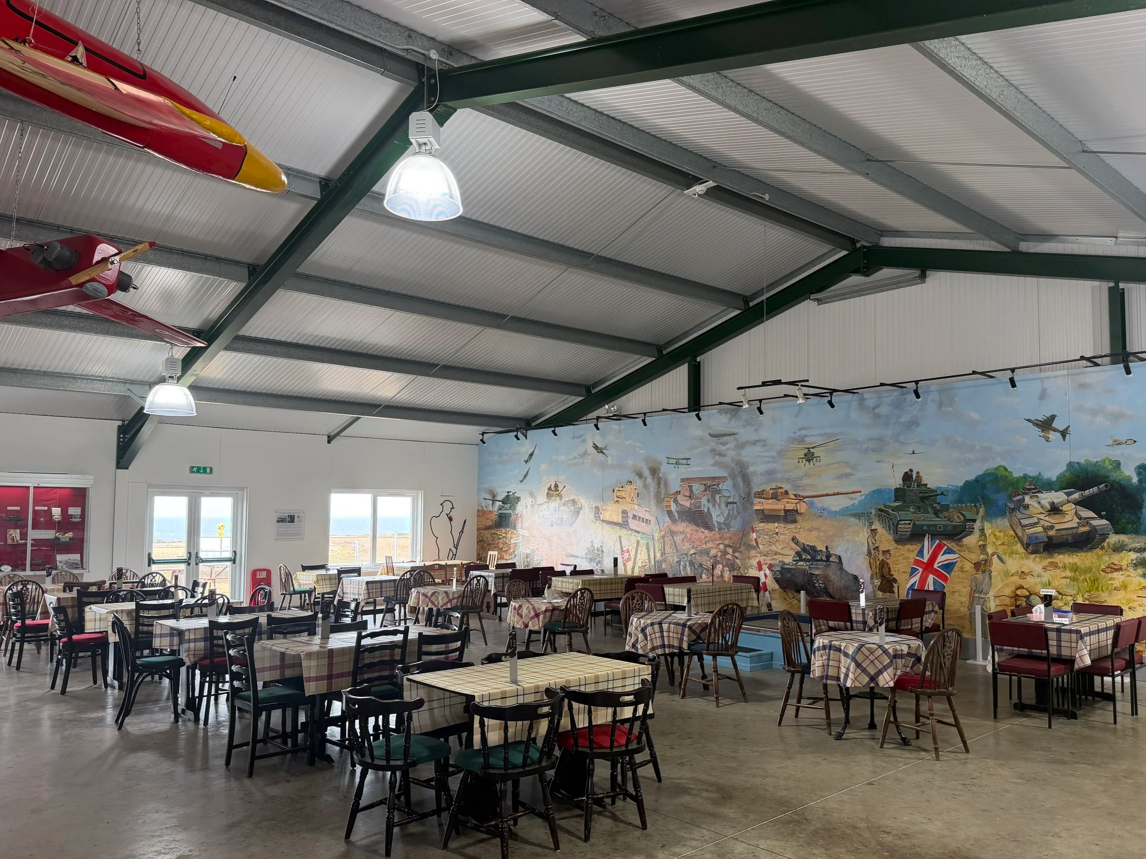 Dining area with tables covered in checkered tablecloths and chairs, a large mural of military tanks and aircraft on the wall, windows letting in natural light, and fire extinguishers and exit signs visible.
